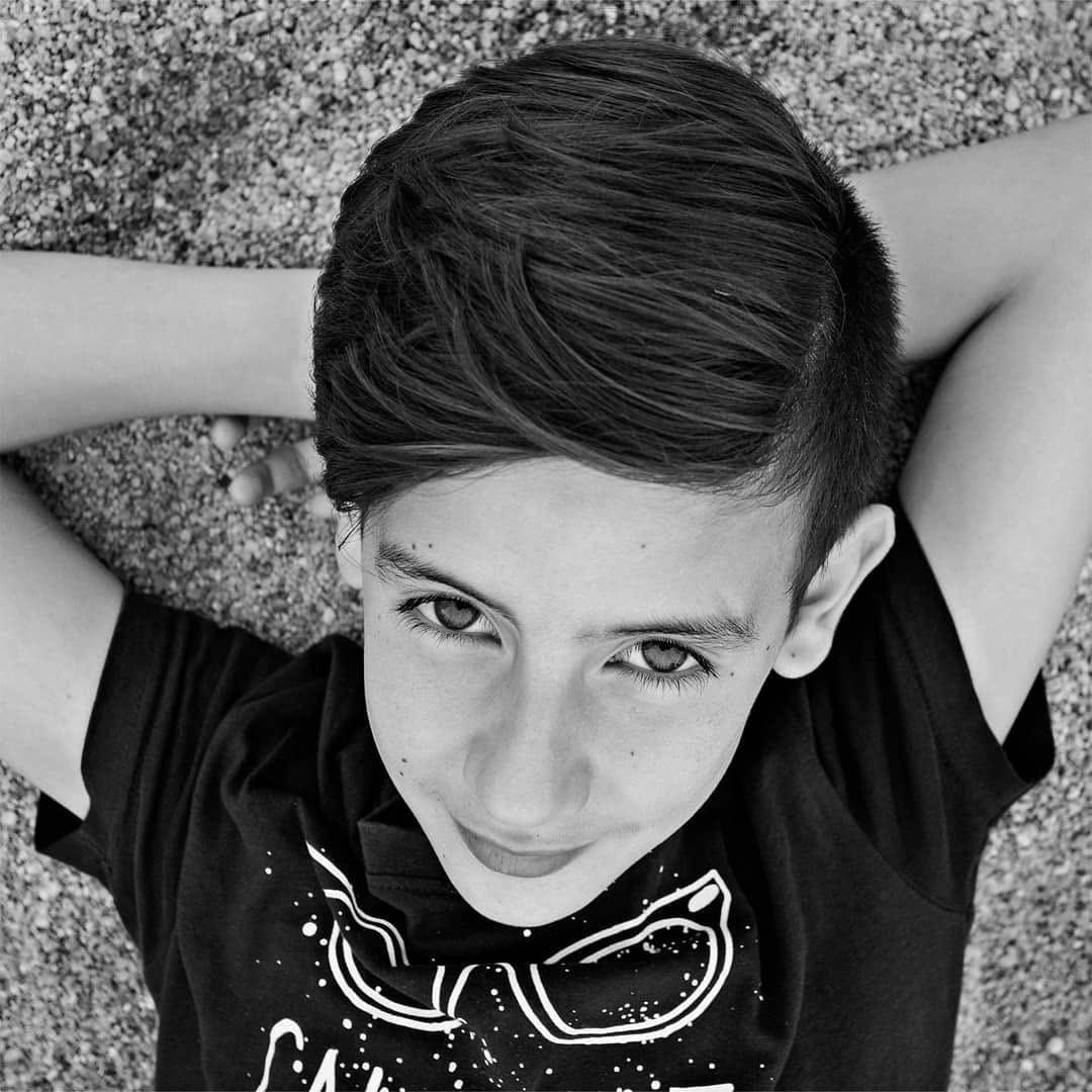 Segunda variante en blanco y negro del mismo niño, ahora con expresión más serena y una sonrisa leve, en la misma pose recostada sobre la superficie granulada.