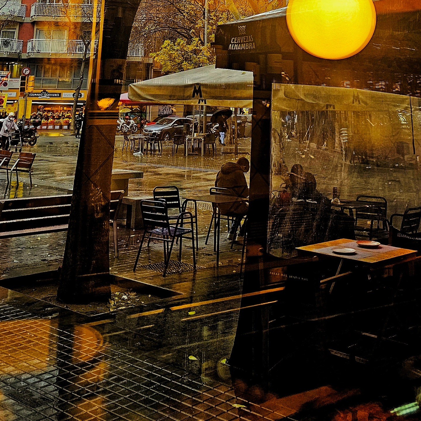 Reflejo invertido de una terraza de bar bajo toldos, con mesas metálicas vacías, un cartel de 'Cervezas Alhambra' y un gran disco de luz amarilla que domina la escena, todo capturado en un…