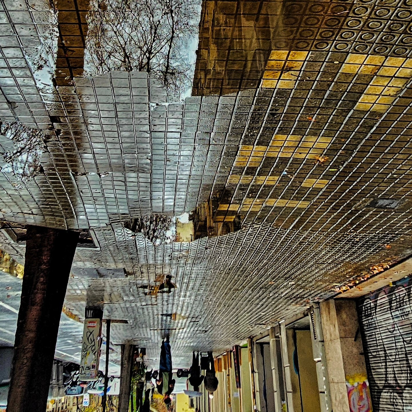 Reflejo invertido de una galería o pasaje con techo de rejilla metálica, persianas cerradas de locales comerciales y graffiti en los pilares, capturado en un charco sobre baldosas amarillas…