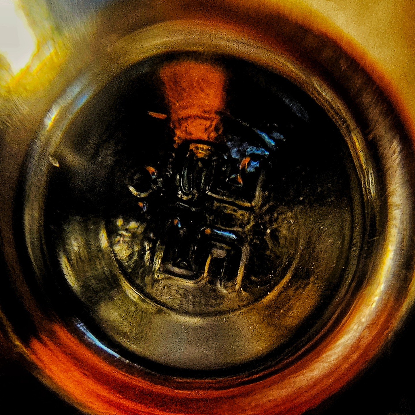 Macrofotografía circular de una taza vista desde arriba, con líquido oscuro y una forma anaranjada borrosa reflejada en el fondo metálico brillante, similar en estilo a la otra imagen de…