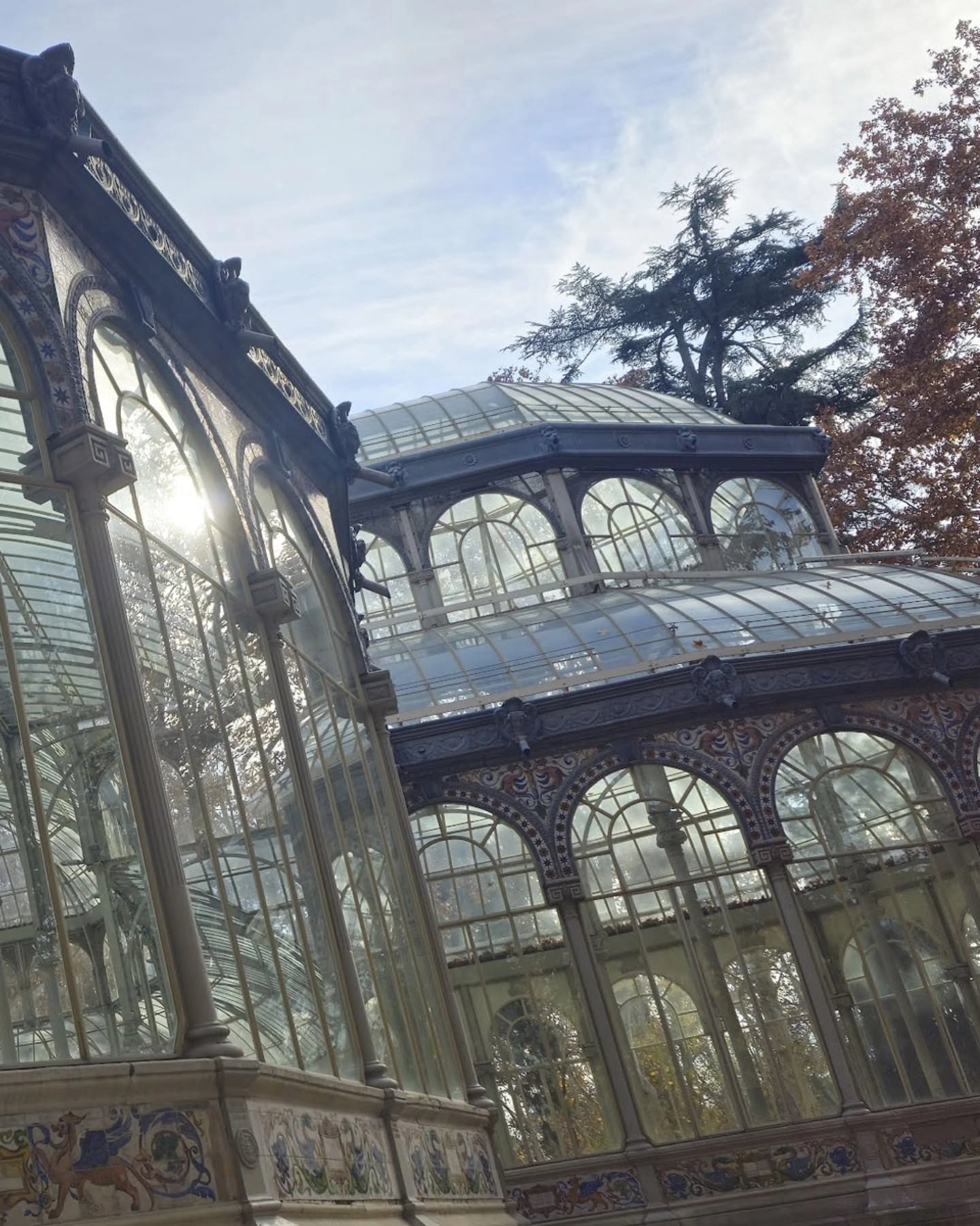 Toma exterior en contrapicado del Palacio de Cristal, con su estructura de hierro y vidrio recortada contra un cielo despejado y un árbol de hojas otoñales a la derecha.