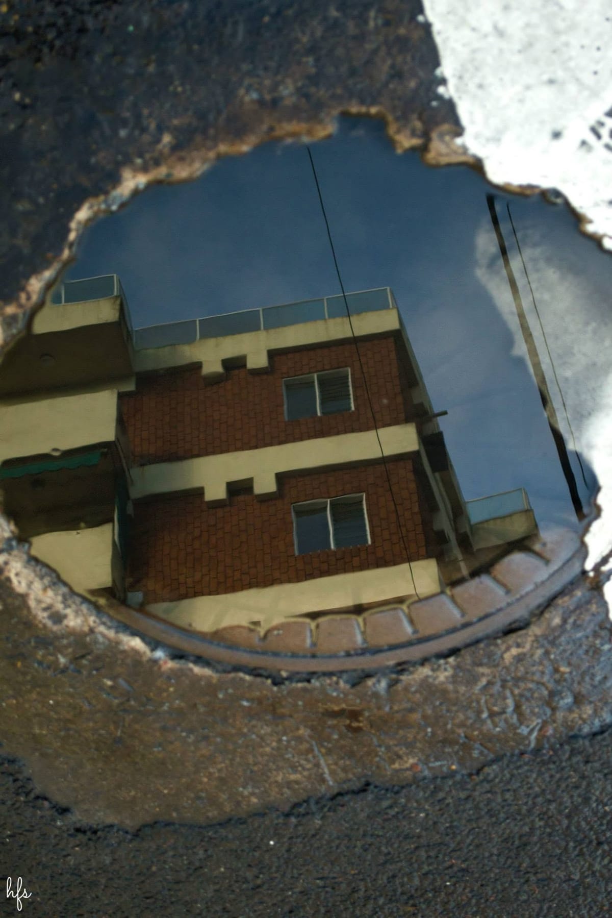 Fotografía a color del reflejo de un edificio de ladrillo visto en un charco de agua sobre el asfalto, con cielo azul y balcones reflejados boca abajo; el borde irregular del charco enmarca…