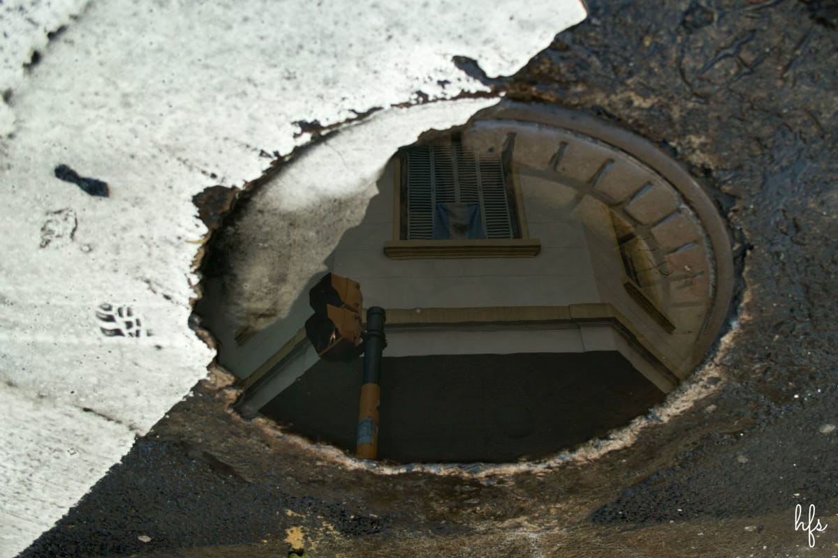 Vista cenital de baldosas de vereda mojadas donde se refleja, distorsionada, la silueta oscura de un semáforo o cartel de señalización colgante.