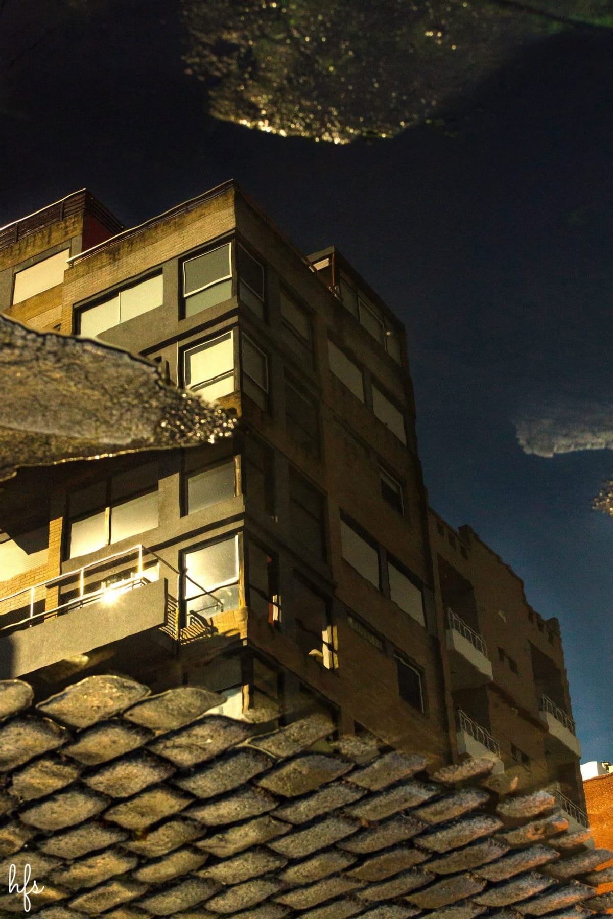 Fotografía a color del reflejo nocturno de un edificio de departamentos iluminado, visto en un charco sobre baldosas de vereda; ventanas encendidas y balcones se reflejan invertidos contra…