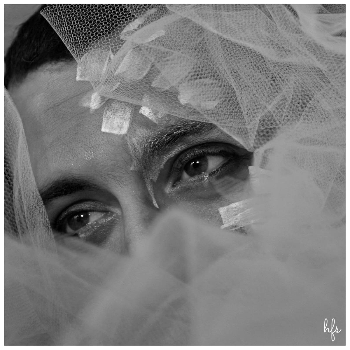 Fotografía macro en blanco y negro de un par de ojos de mirada fija, cubiertos parcialmente por capas de tul en diagonal con textura de panal; composición muy cerrada y de alto contraste.