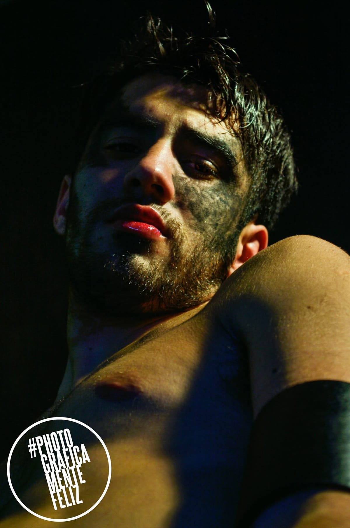 Retrato dramático de un hombre con el pelo mojado y el rostro parcialmente cubierto de sombra y suciedad, labios brillantes, torso desnudo apoyado sobre la propia rodilla, iluminación…