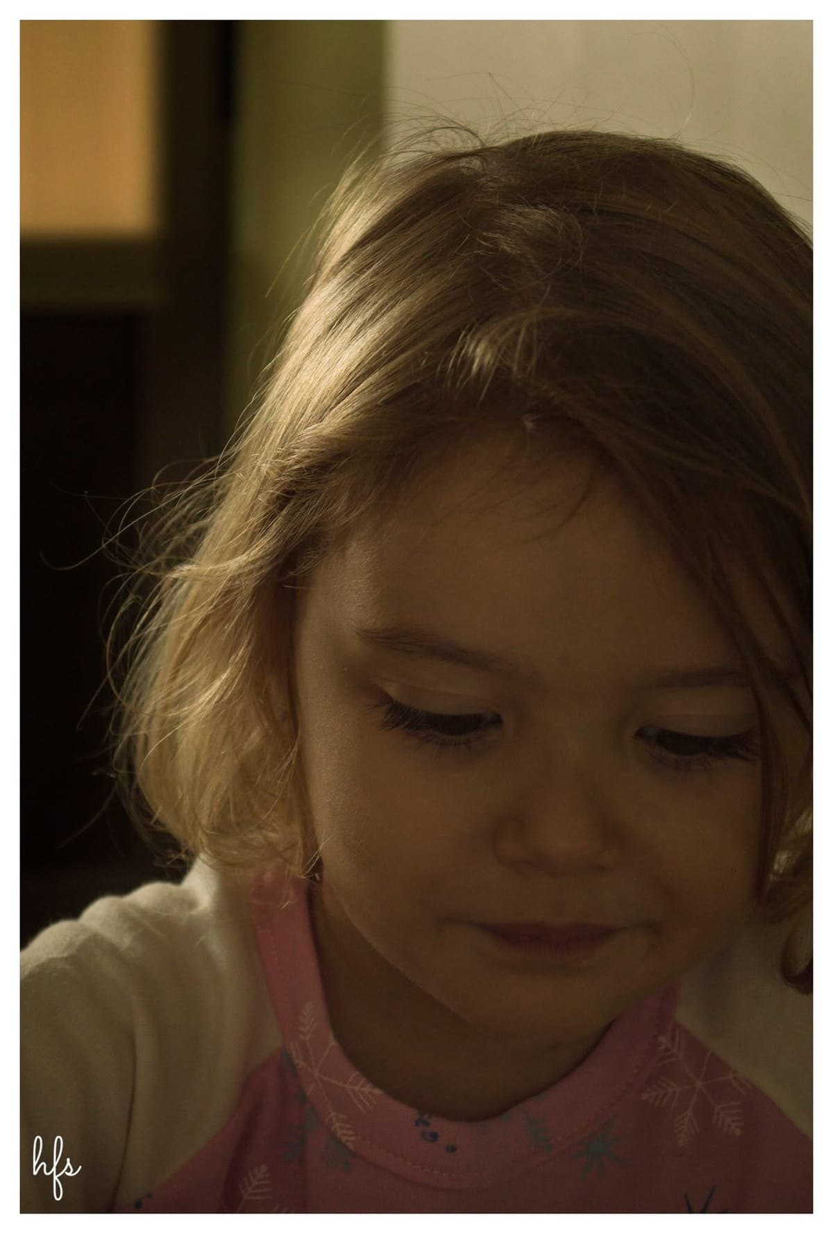 Retrato a color en primer plano de una niña pequeña de pelo castaño claro iluminado por luz cálida lateral, mirando hacia abajo con una leve sonrisa; viste una prenda rosa con estampado de…