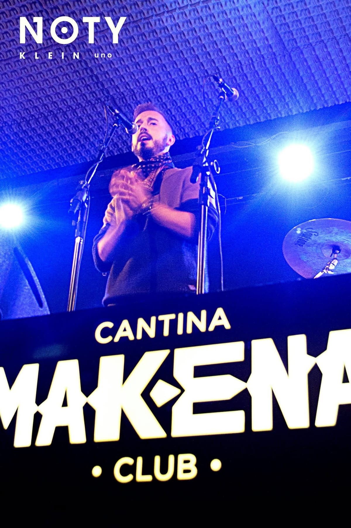 El cantante principal, visto desde abajo, canta con las manos cruzadas sobre el pecho y pañuelo al cuello, mientras el cartel "Cantina Mak-ena Club" ocupa la parte inferior del encuadre.