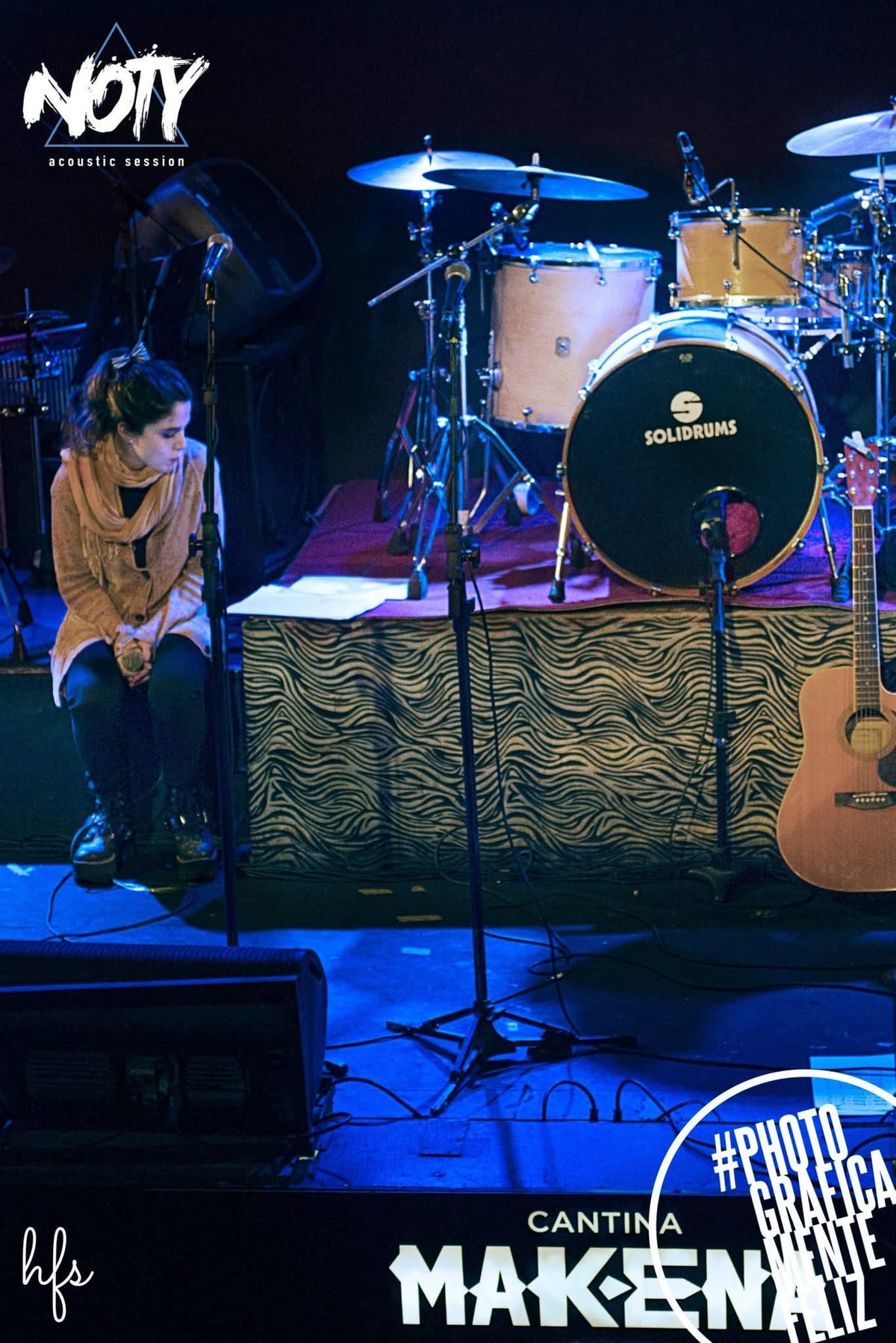 El cantante, sentado en un banco alto con sombrero y poncho, canta con las manos juntas frente al pecho, corista y amplificador rosa desenfocados detrás.