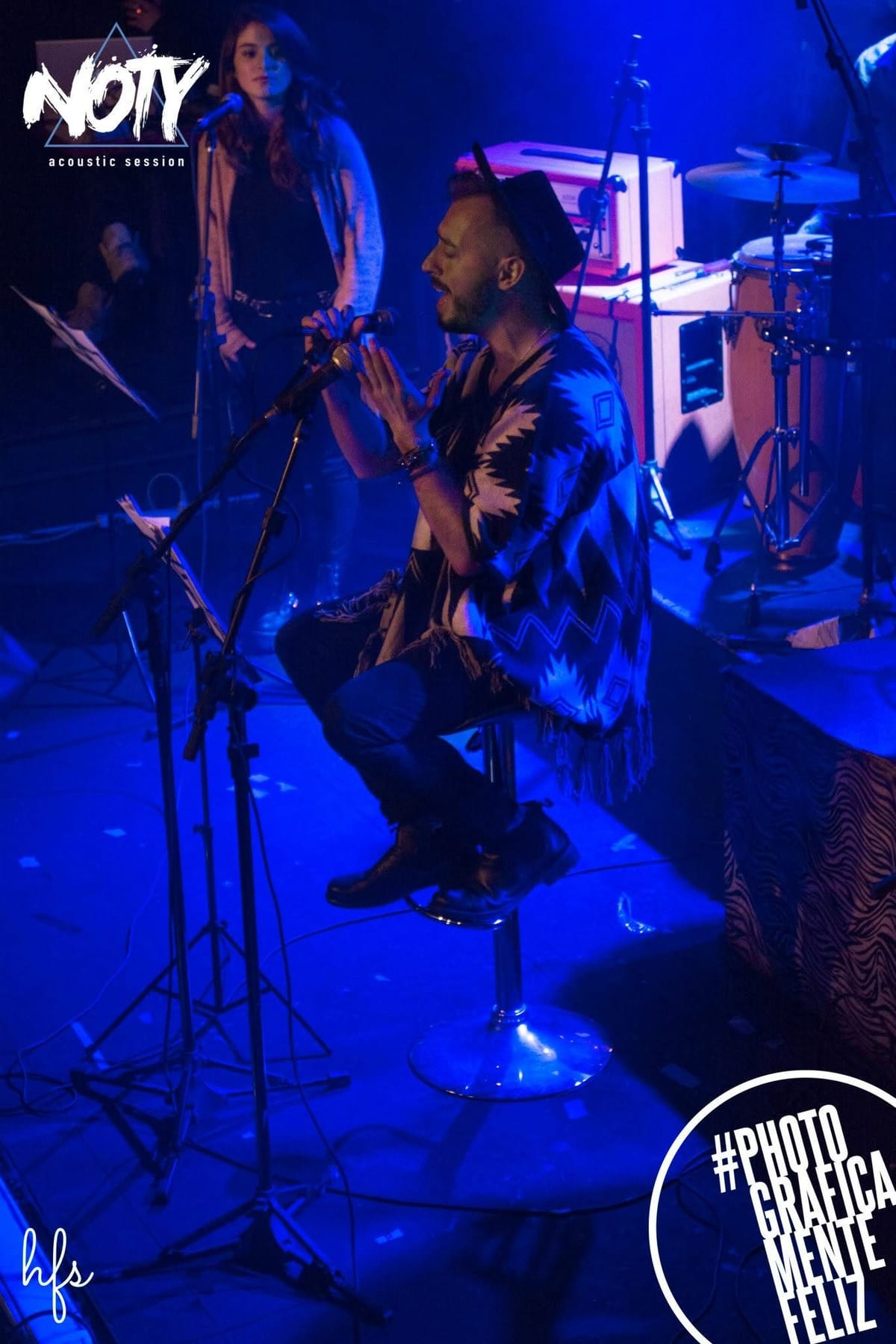 Baterista de perfil en primer plano borroso por el movimiento de la cámara, con dos compañeros de banda desenfocados tocando guitarra y cantando al fondo bajo luces violetas.