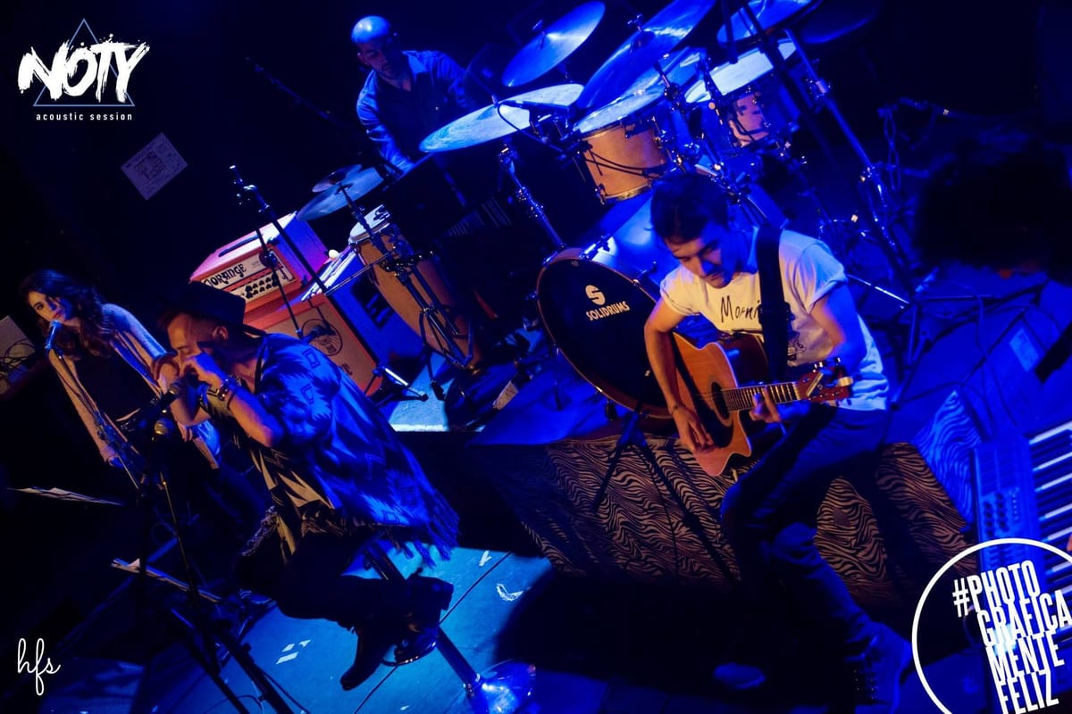 Plano amplio de la banda en acción: una cantante y el cantante principal al frente sobre los micrófonos, un baterista y un guitarrista tocando detrás bajo luz azul intensa.