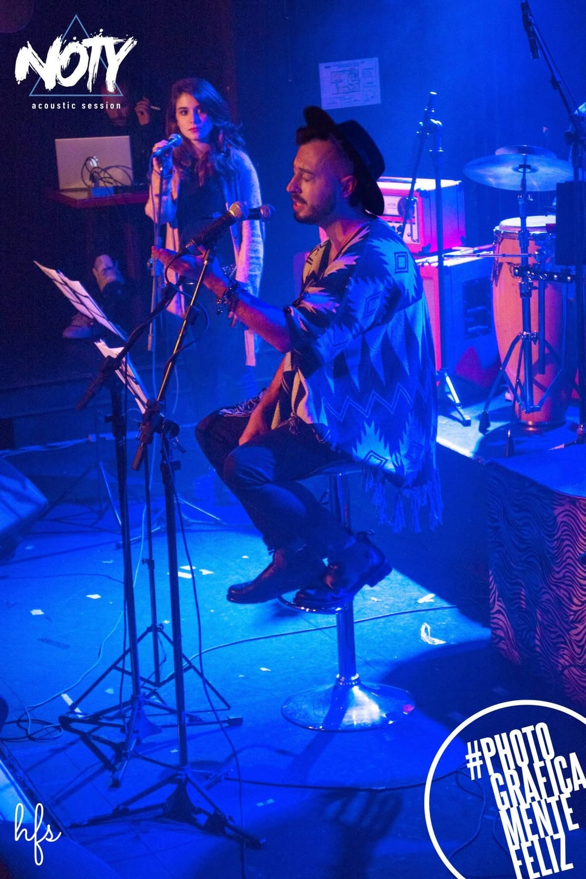 Fotografía a color de un cantante sentado en un banco alto, con poncho y sombrero, cantando con los ojos cerrados; detrás, una corista canta también en su propio micrófono, bajo una intensa…