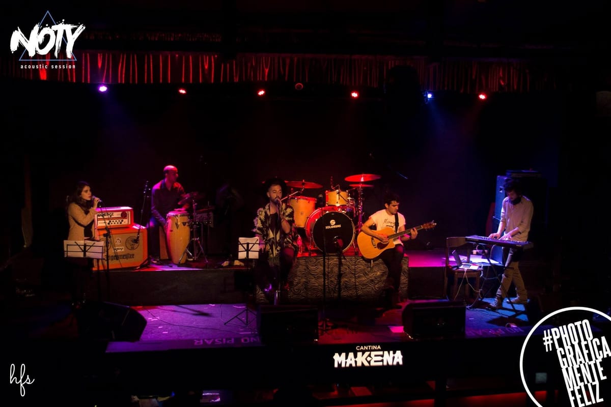 El cantante mira hacia arriba, con la boca abierta cantando, iluminado por un contraluz azul intenso que recorta su silueta contra el cartel «Cantina Makena Club» en letras blancas sobre…
