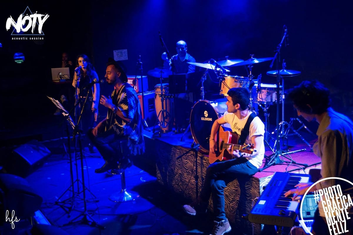 Guitarrista de pie afinando o tocando su instrumento en el borde del escenario, con otro músico sentado tocando la guitarra a su lado, bajo un intenso baño de luz violeta y azul.