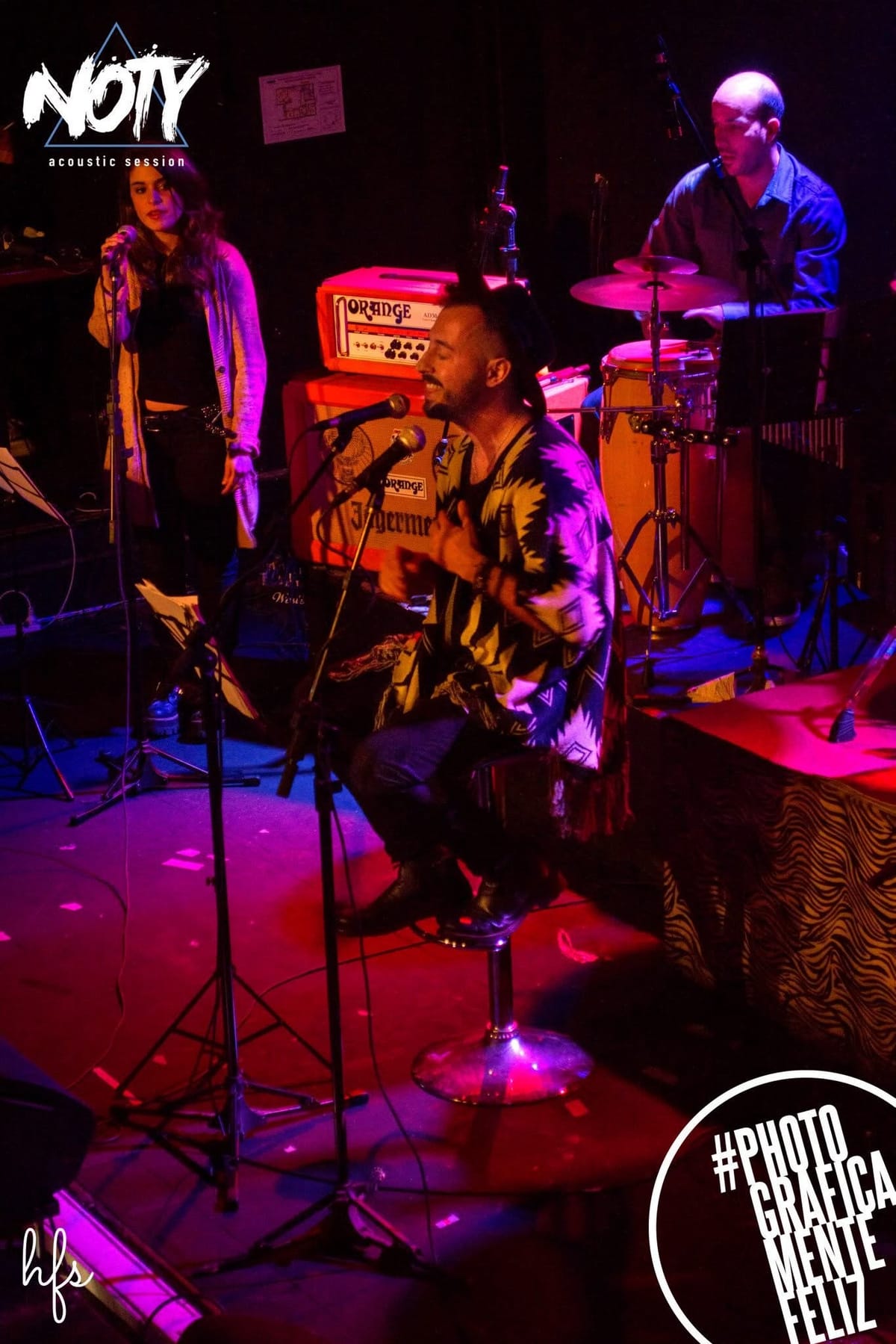 El cantante con sombrero y poncho canta junto a una corista, ambos frente a sus micrófonos, con amplificadores y congas iluminados en tonos rosados detrás.