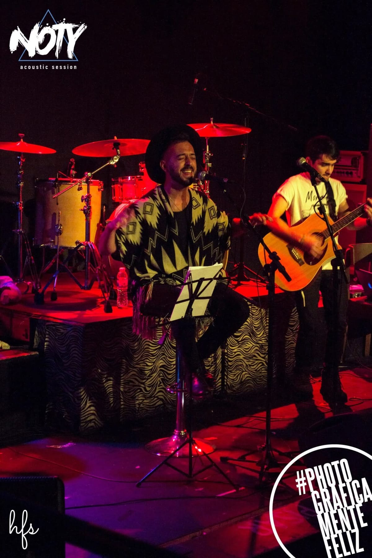 Silueta de un cantante con sombrero aplaudiendo o gesticulando entre humo y luz azul intensa sobre el escenario, con la batería y platillos detrás; el cartel iluminado 'CANTINA MAKENA CLUB'…