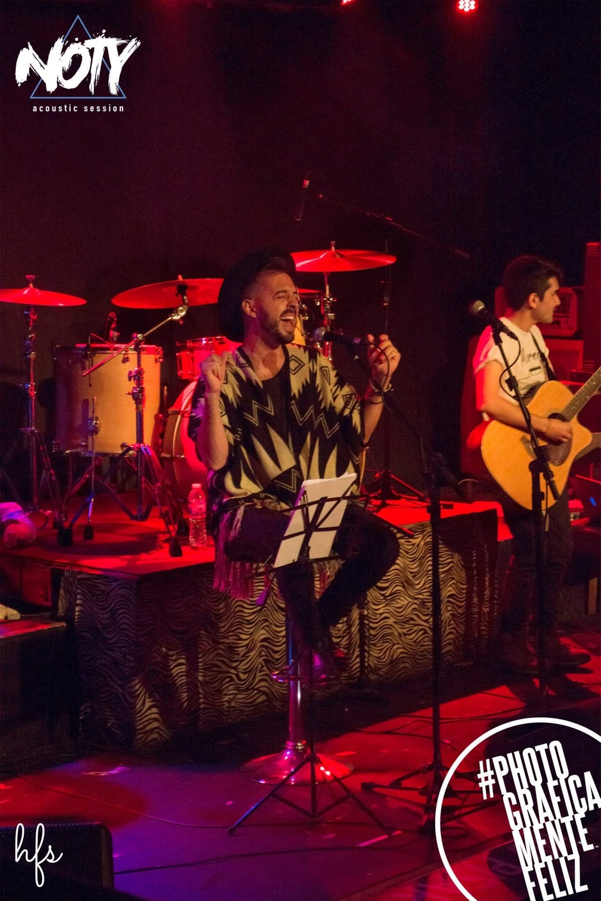 Corista sentada con abrigo mostaza y bufanda cantando junto a un atril de partituras, batería y guitarra acústica apoyada visible al costado del escenario en luz azul.