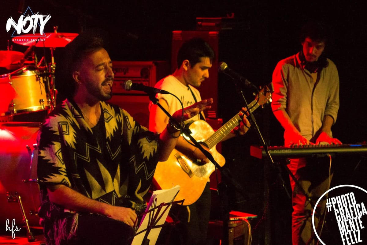 Cantante con poncho estampado sentado en un taburete cantando ante el micrófono, junto a un guitarrista de remera clara tocando guitarra acústica, ambos bañados en luz roja cálida de…