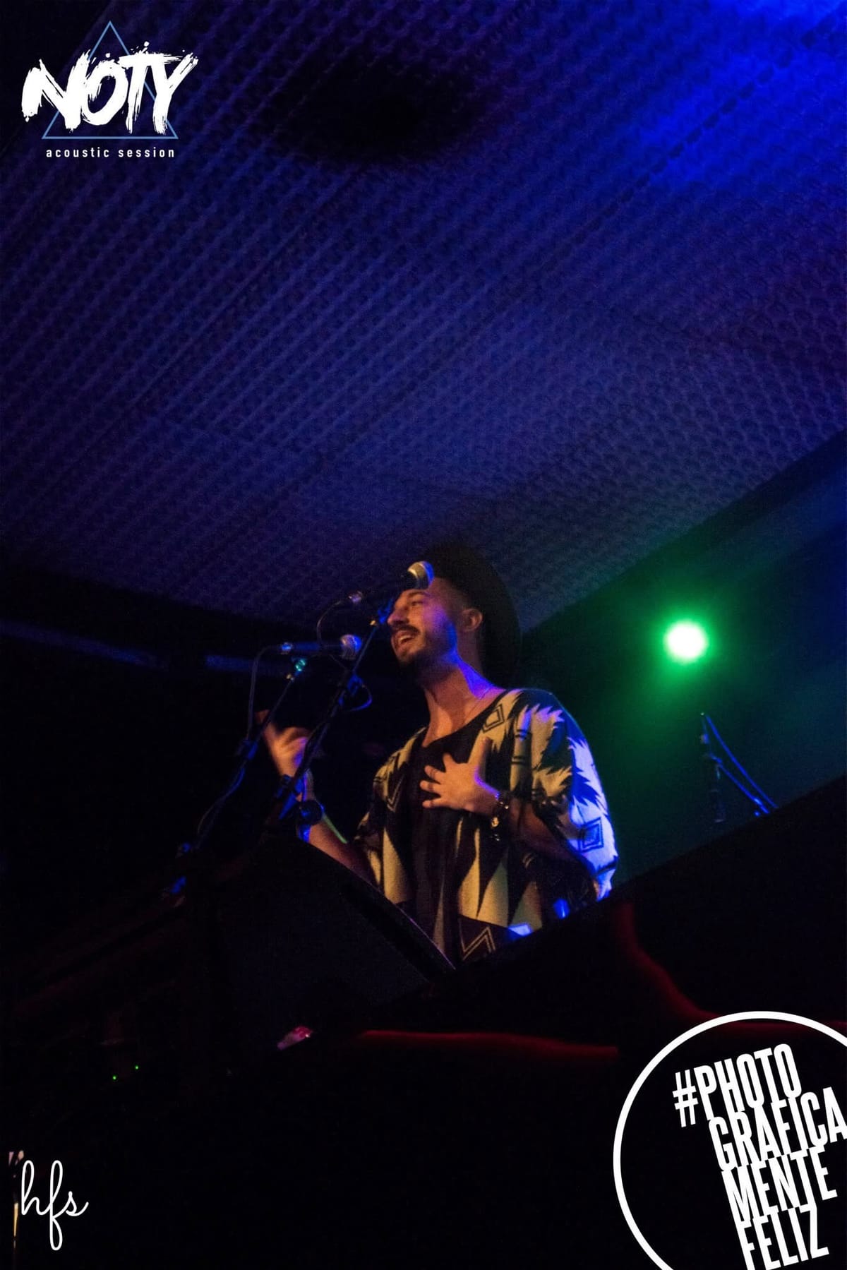 El cantante, con sombrero, canta con expresión intensa y una mano sobre el pecho, bajo un foco verde que contrasta con el ambiente azulado del resto del escenario.