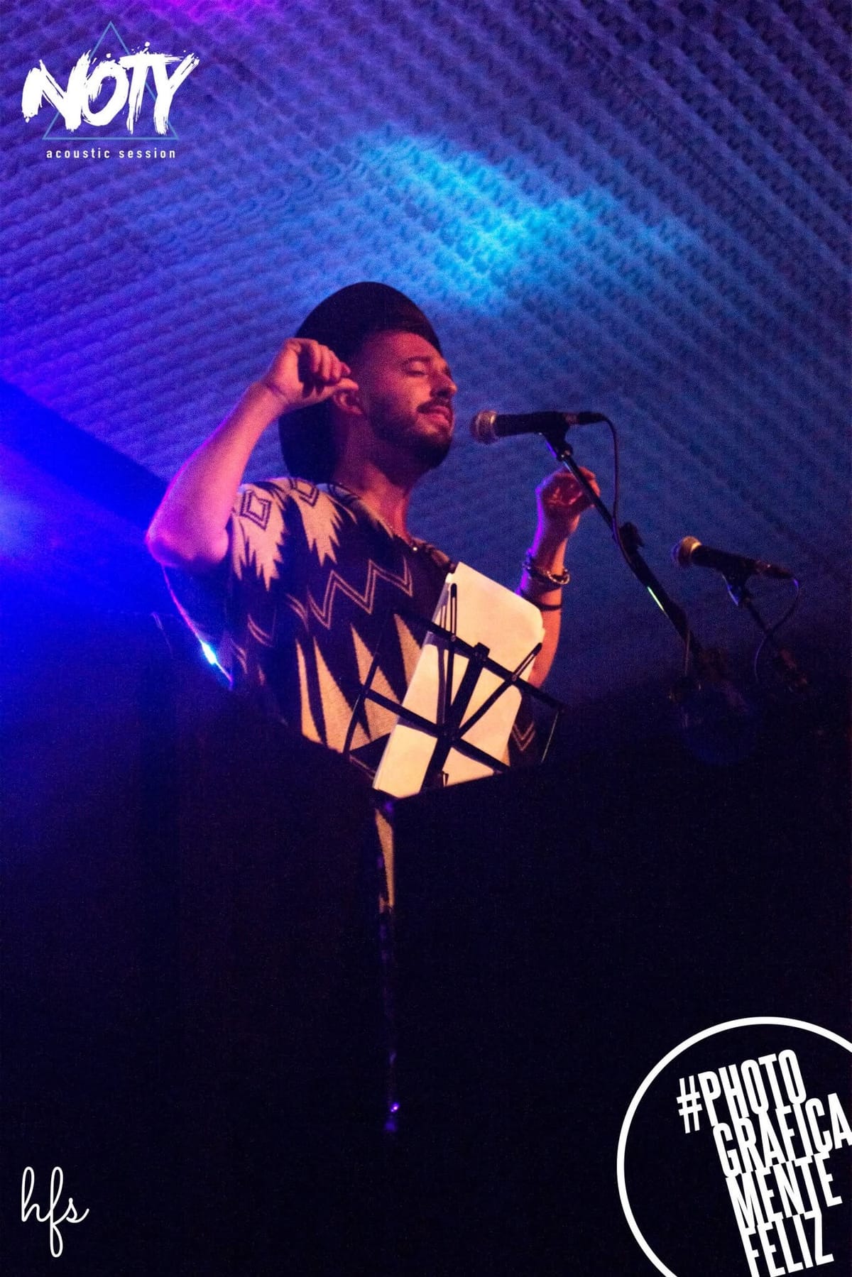 Fotografía a color de un cantante con gorra hacia atrás y remera estampada, con un brazo levantado junto a la cabeza y el otro extendido hacia el micrófono, bajo una intensa luz violeta y…