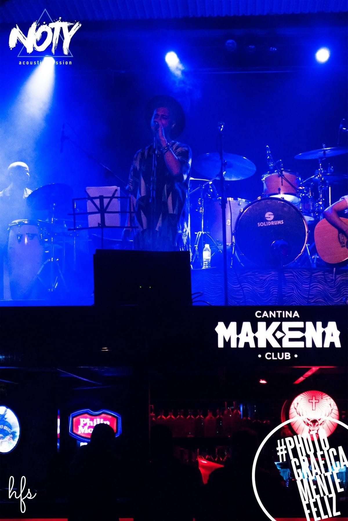 Vista amplia del escenario con varios músicos: una corista cantando junto a un micrófono, el cantante principal con poncho, un guitarrista sentado y un baterista al fondo, todos bajo…
