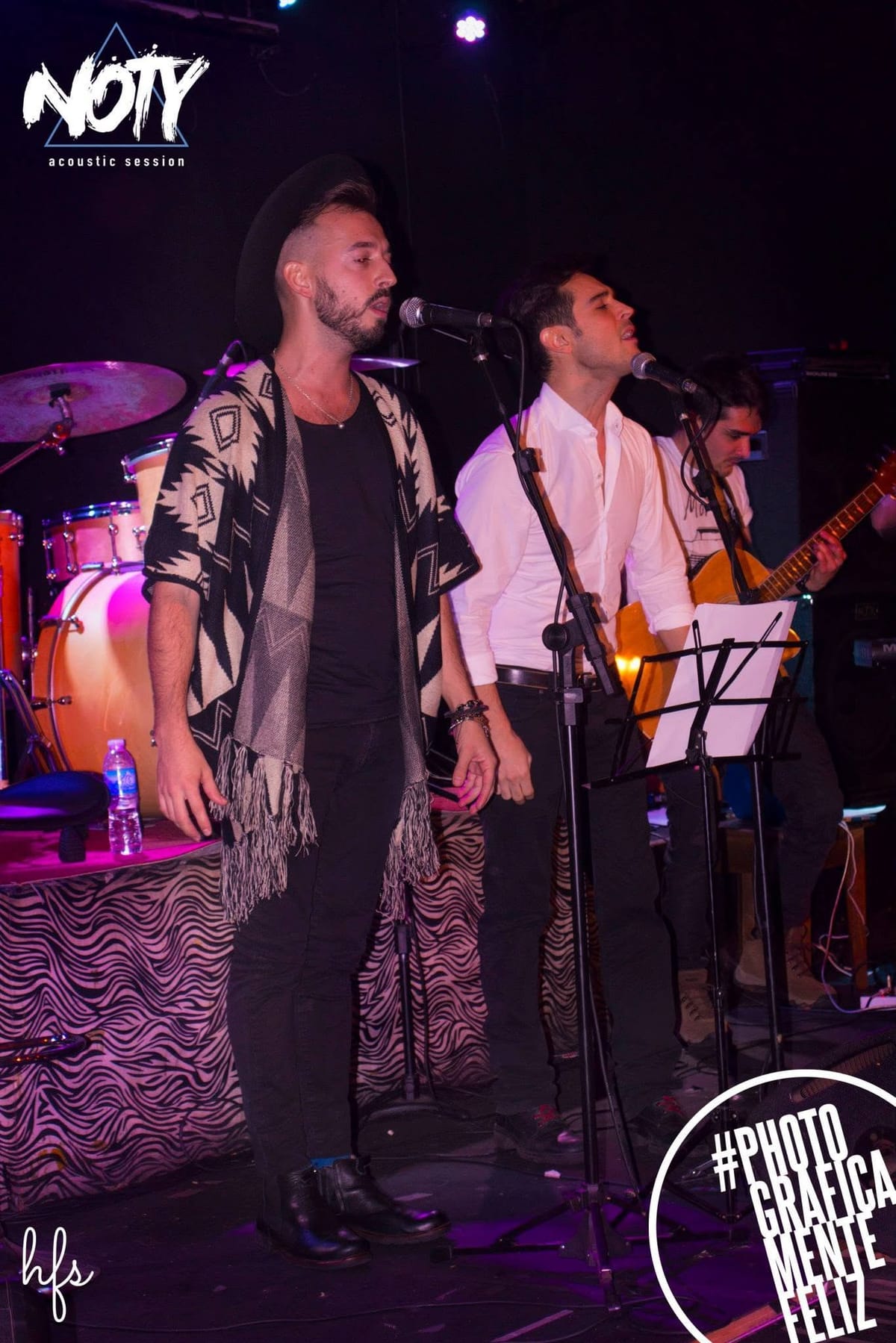 Tres músicos cantando juntos frente a sus micrófonos: uno con sombrero y poncho, otro de camisa blanca, un tercero al fondo tocando guitarra, con atril de partituras entre ellos.