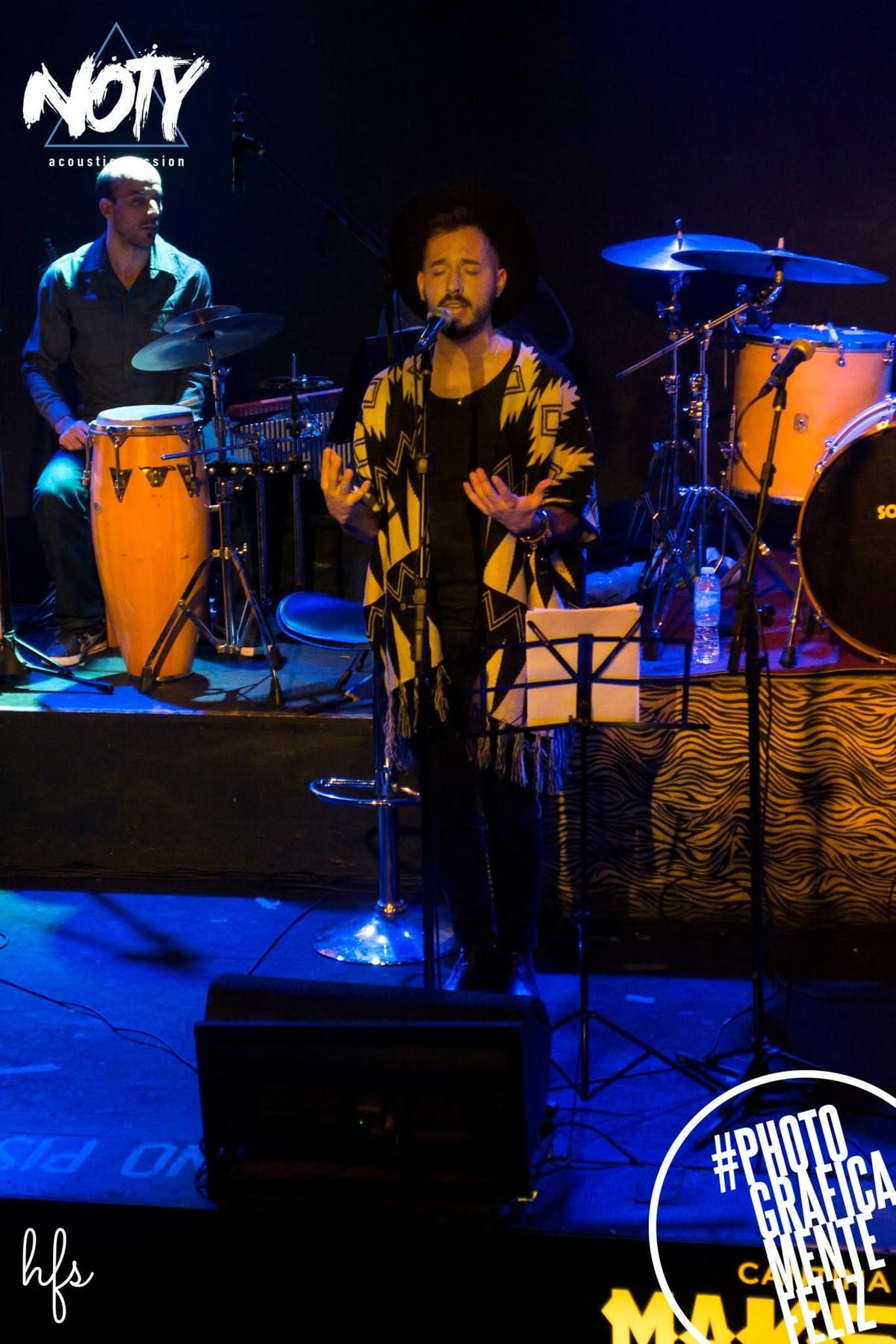 El cantante con sombrero y poncho abre las manos en gesto expresivo mientras canta, con un percusionista tocando congas detrás y la batería anaranjada iluminada al fondo.