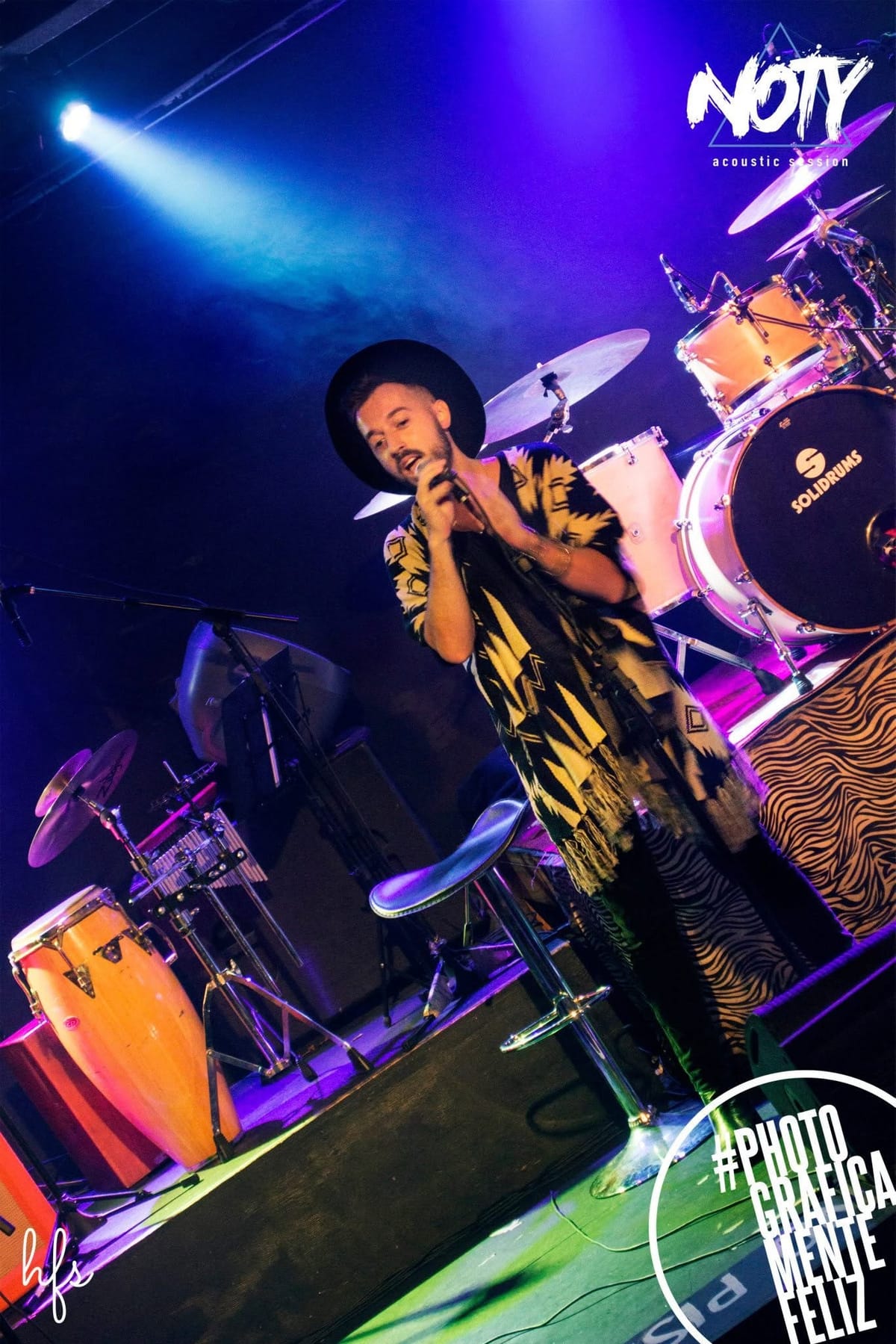 Cantante con sombrero negro de ala ancha y poncho estampado en blanco y negro, cantando al micrófono sobre el escenario, batería y platillos iluminados en azul detrás, banqueta vacía a un…