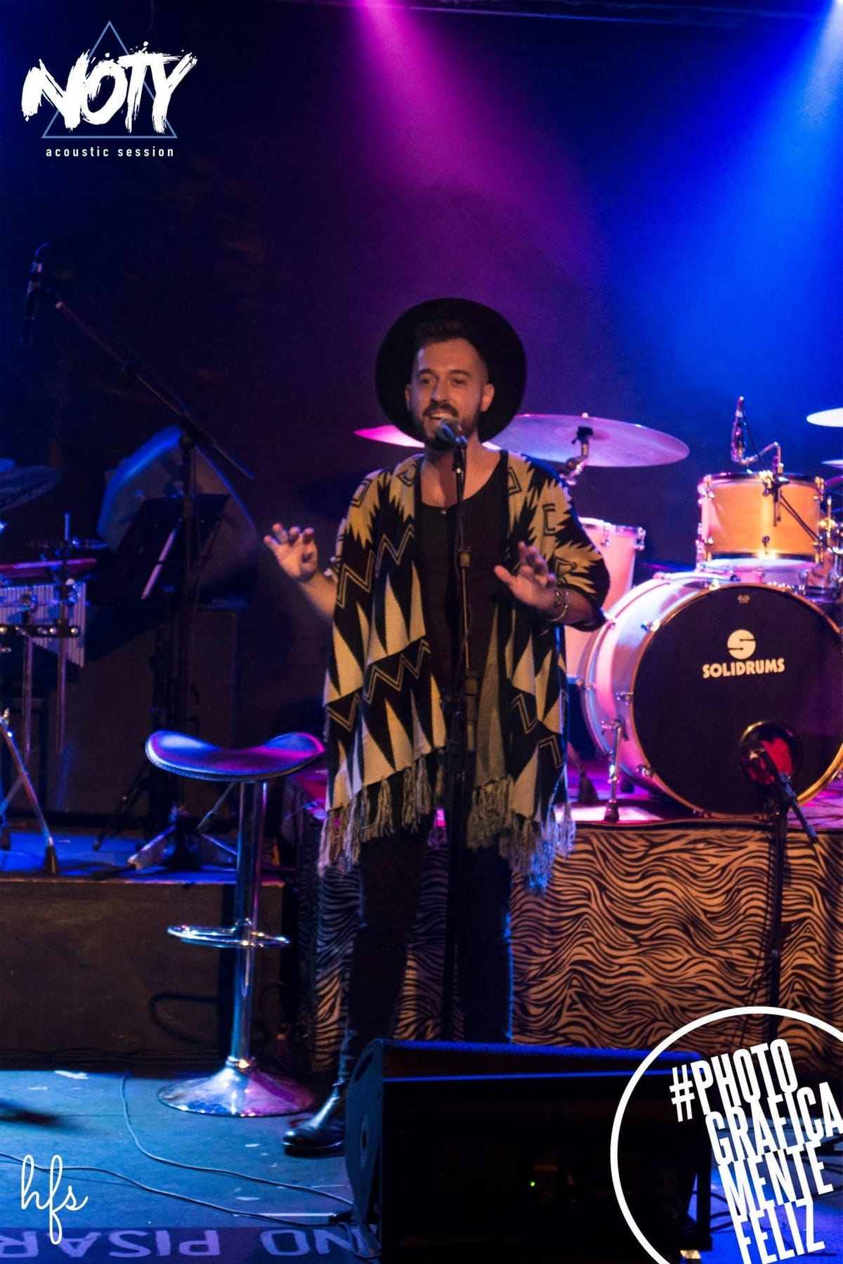 Fotografía a color de un cantante con sombrero negro de ala y poncho estampado en blanco y negro, cantando con los brazos abiertos frente al micrófono; detrás, una batería con parche…