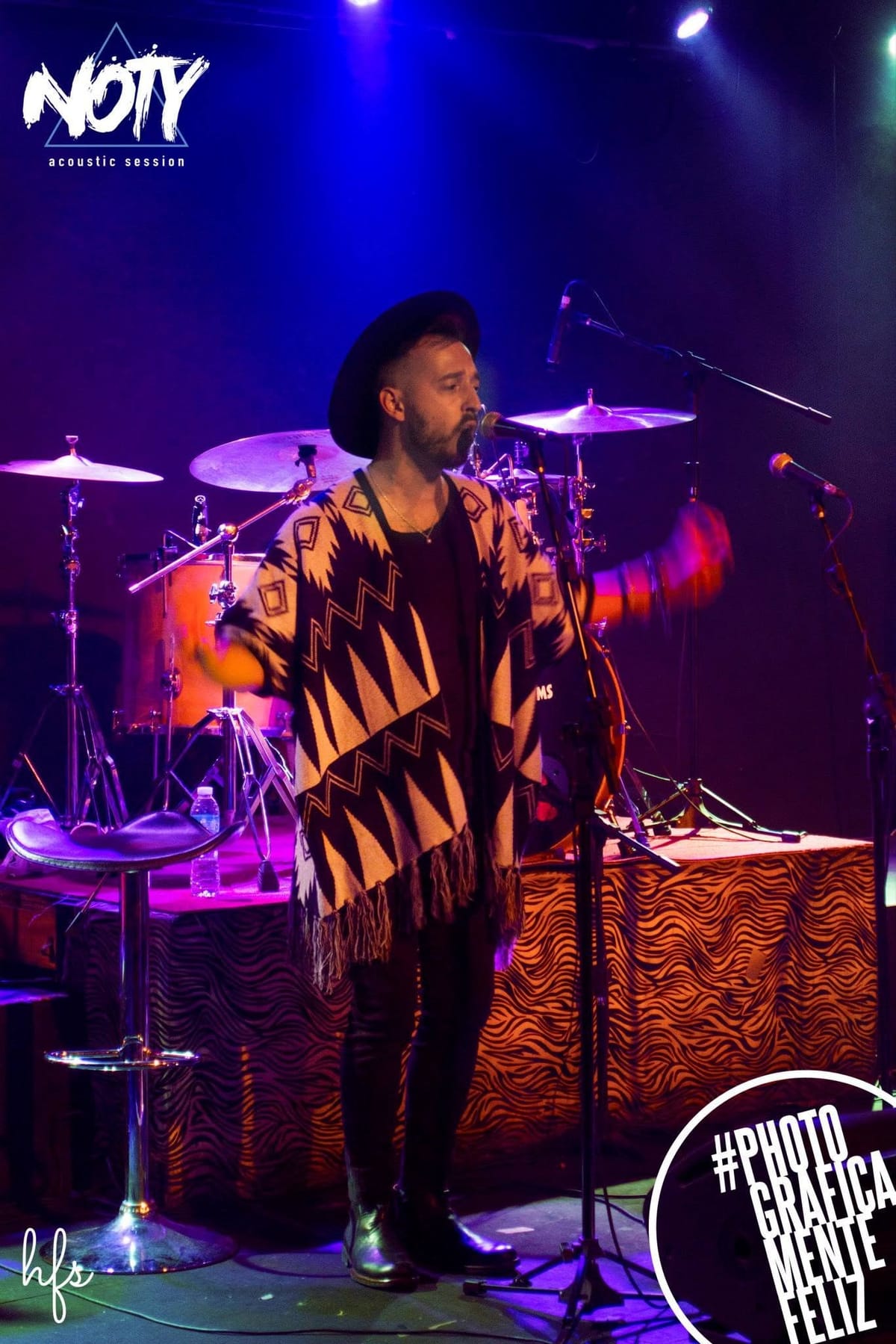 Plano general del cantante de pie cantando al micrófono, escenario con batería naranja y amplificadores Orange, cartel "Cantina Mak-ena Club" al frente del escenario, luces azules…