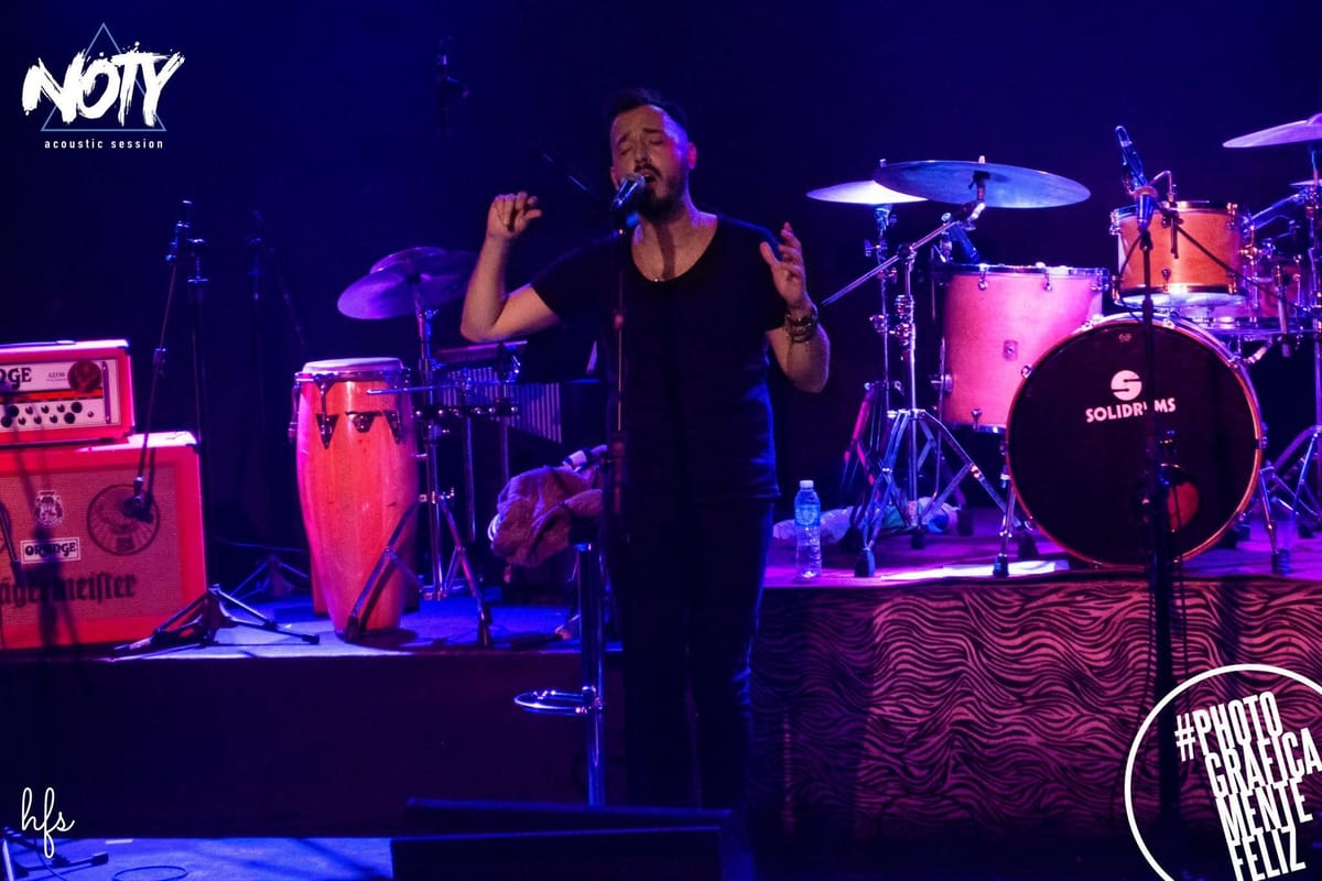 Dos cantantes comparten el frente del escenario, uno con poncho estampado y otro con camisa rosa, ambos cantando a micrófono bajo una luz azul intensa que recorta sus siluetas contra la…