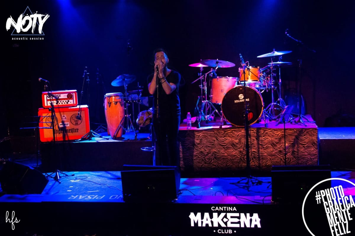 Plano general del cantante solo en escena, cabeza inclinada y mano cerca de la boca sobre el micrófono, con la batería naranja "Solidrums", congas y el cartel "Cantina Mak-ena Club"…