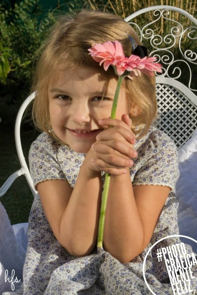 Primer plano de una niña sonriente sosteniendo con ambas manos una gran flor rosa (gerbera) que tapa parcialmente su rostro, sentada en la misma silla de hierro blanco del jardín.
