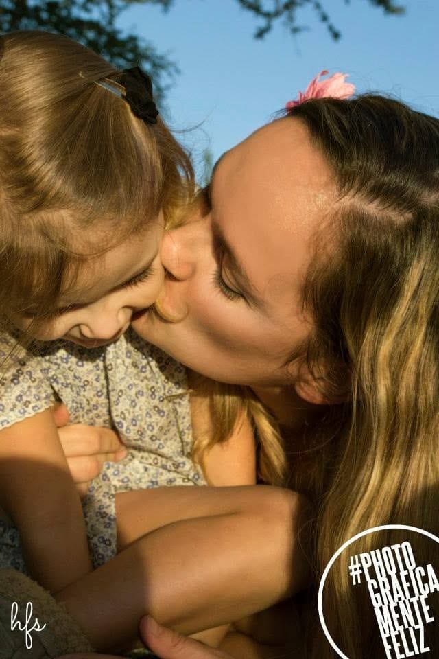 Fotografía a color de una mujer con una flor rosa en el pelo besando la mejilla de una niña pequeña que sonríe con los ojos cerrados, ambas abrazadas bajo un cielo despejado y ramas de árbol.