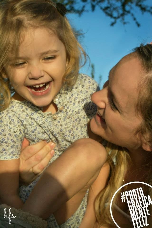 Una niña con vestido floral ríe a carcajadas tomada de la mano de su madre, quien la mira con ternura desde un costado.