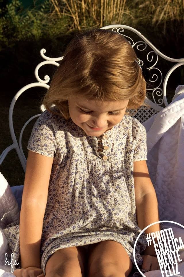 Niña pequeña de pelo castaño claro y vestido floreado, sentada en una silla de hierro forjado blanco en un jardín, mirando hacia abajo con una sonrisa tímida bajo luz cálida de atardecer.