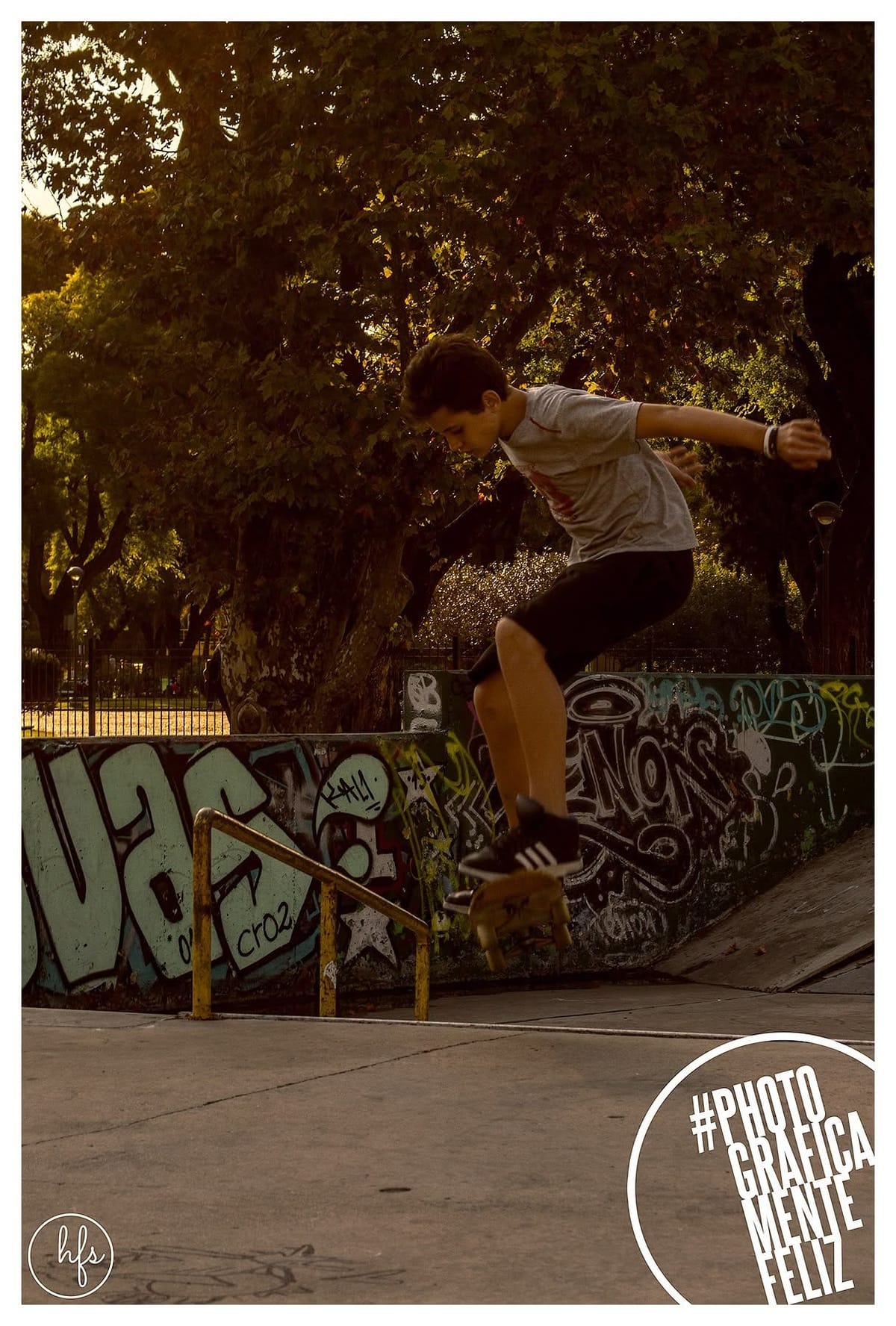 Skater en pleno salto (ollie) con la tabla despegada del piso, remera clara y short oscuro, sobre un fondo de muros cubiertos de grafitis y una baranda amarilla, bajo luz cálida de…