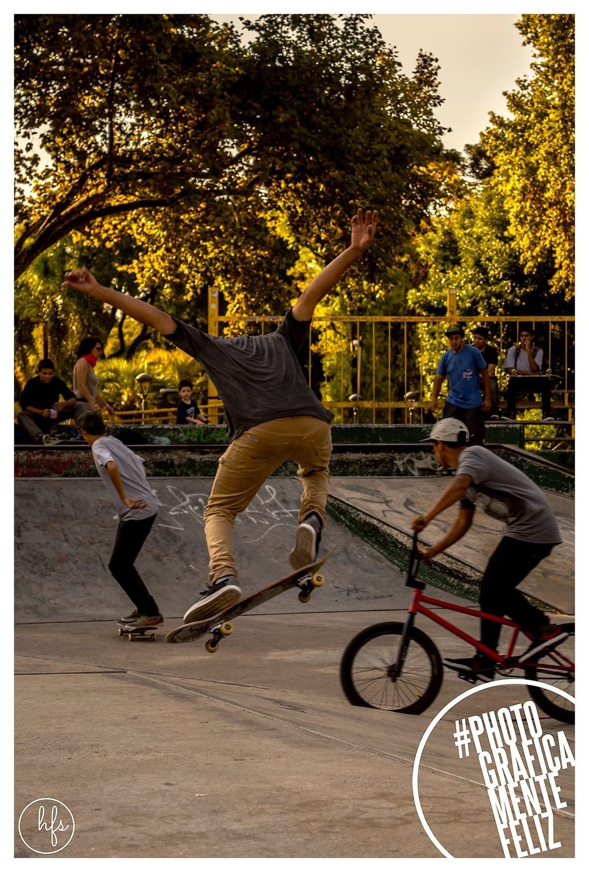 Escena de acción con tres personas simultáneas: un skater salta con los brazos en alto, otro patina hacia la izquierda y un ciclista de BMX avanza en primer plano; fondo de árboles…