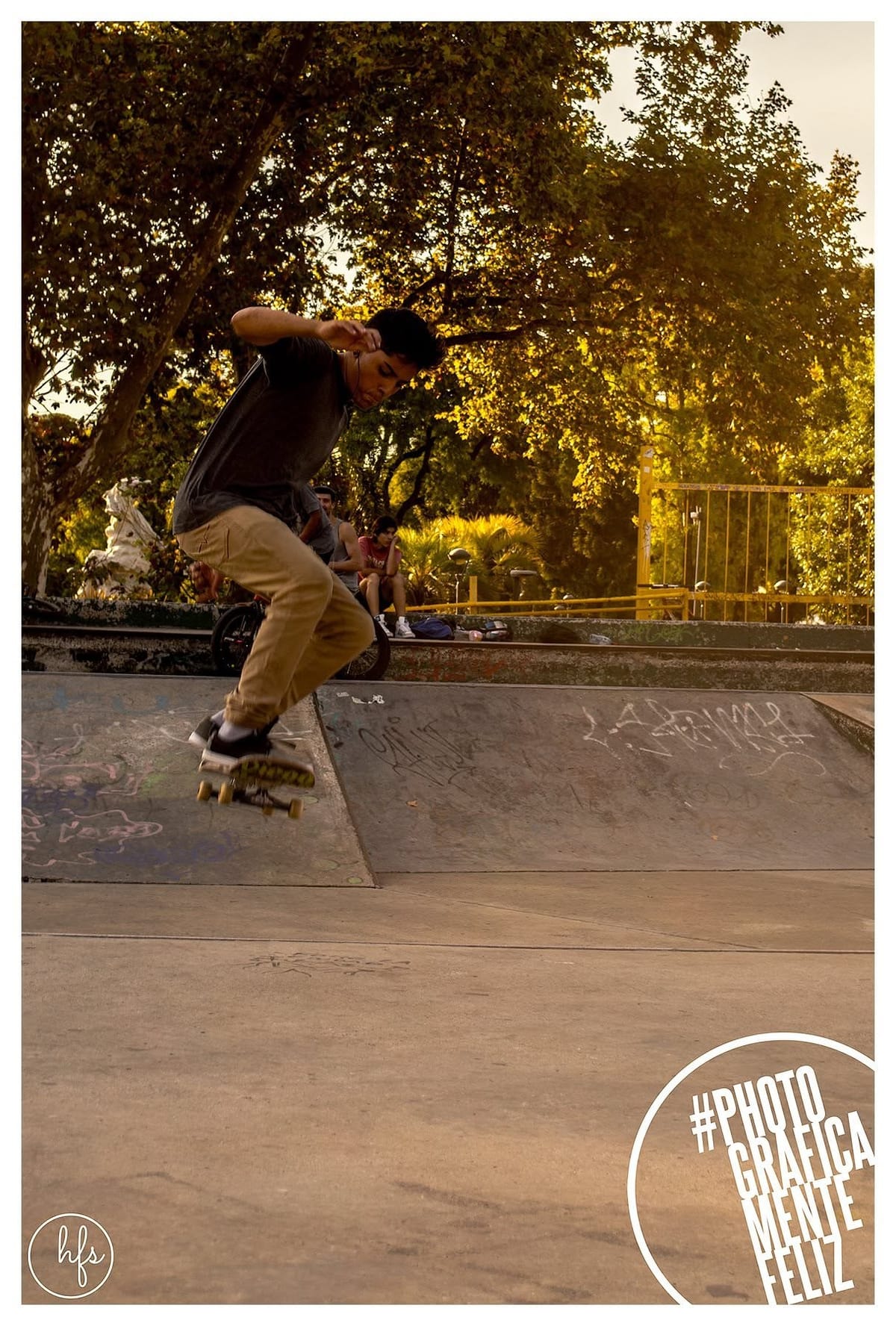 Fotografía a color de un skater saltando desde un borde de cemento grafiteado, con los brazos extendidos en pleno movimiento; al fondo, árboles iluminados por luz de atardecer y otras…