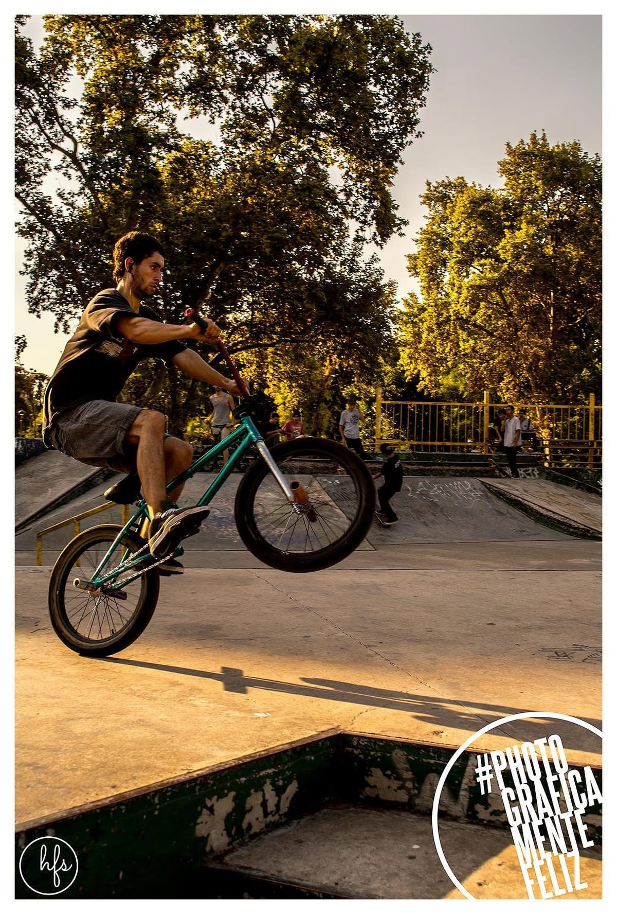 Un ciclista de BMX con remera oscura ejecuta un salto con la bicicleta verde en ángulo, las ruedas despegadas del piso, mientras otros jóvenes observan sentados en la rampa al fondo.