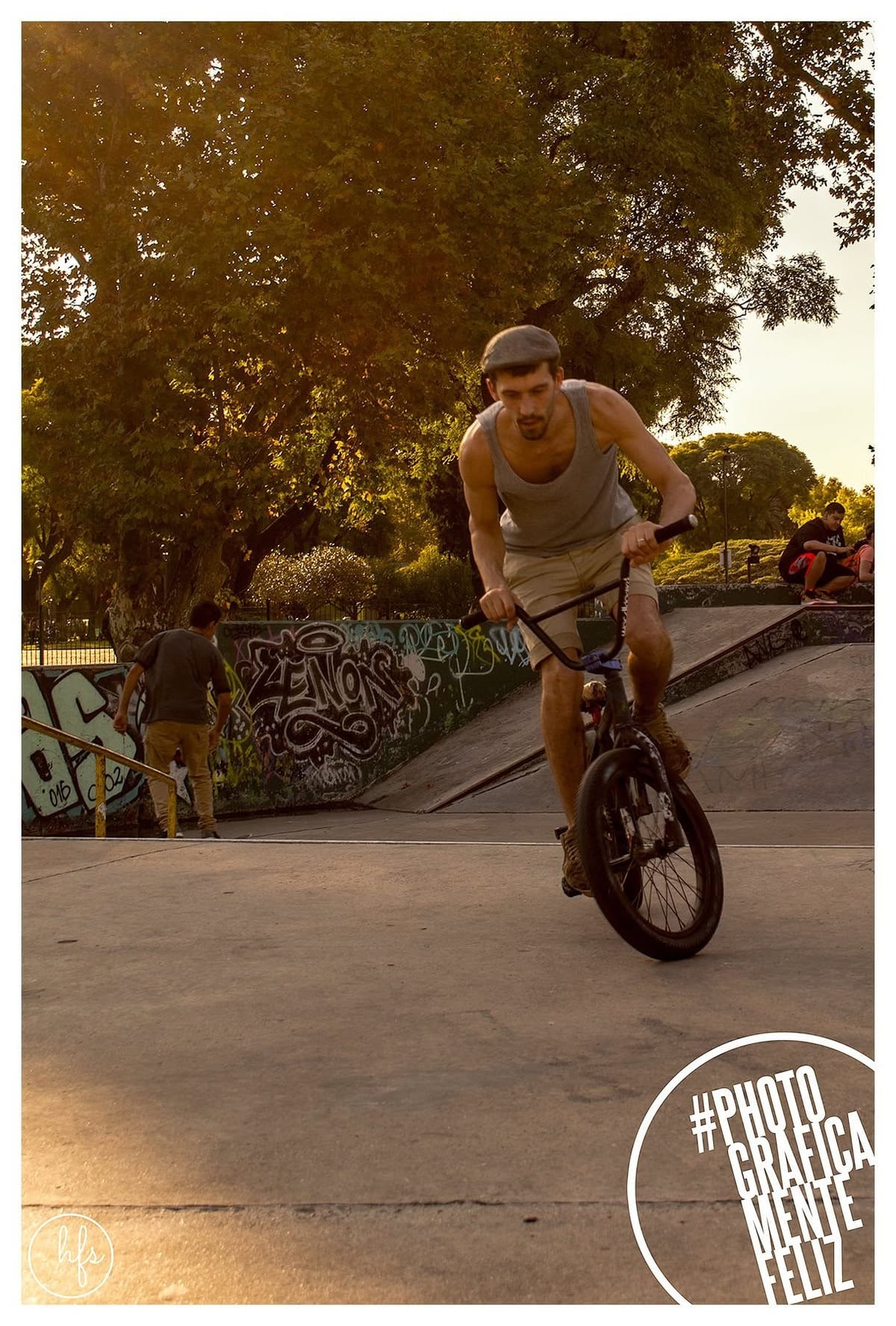 Fotografía a color de un hombre con gorra plana haciendo un wheelie en BMX dentro de una pista de skate, con luz dorada de atardecer entre los árboles y un muro grafiteado al fondo.