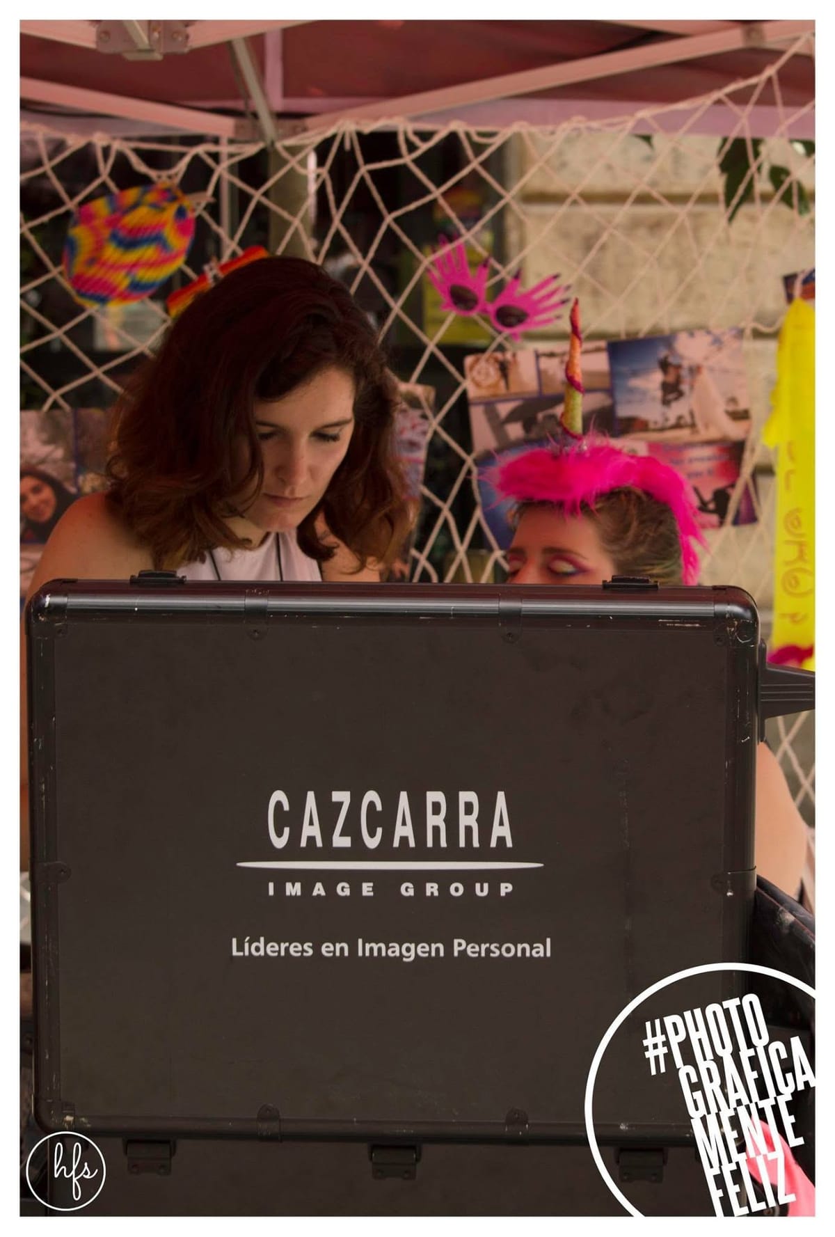 Escena de backstage: una mujer de pelo castaño maquilla a otra que lleva un cuerno de unicornio y plumas fucsia en la cabeza, tras un maletín negro con el logo "CAZCARRA Image Group"; al…