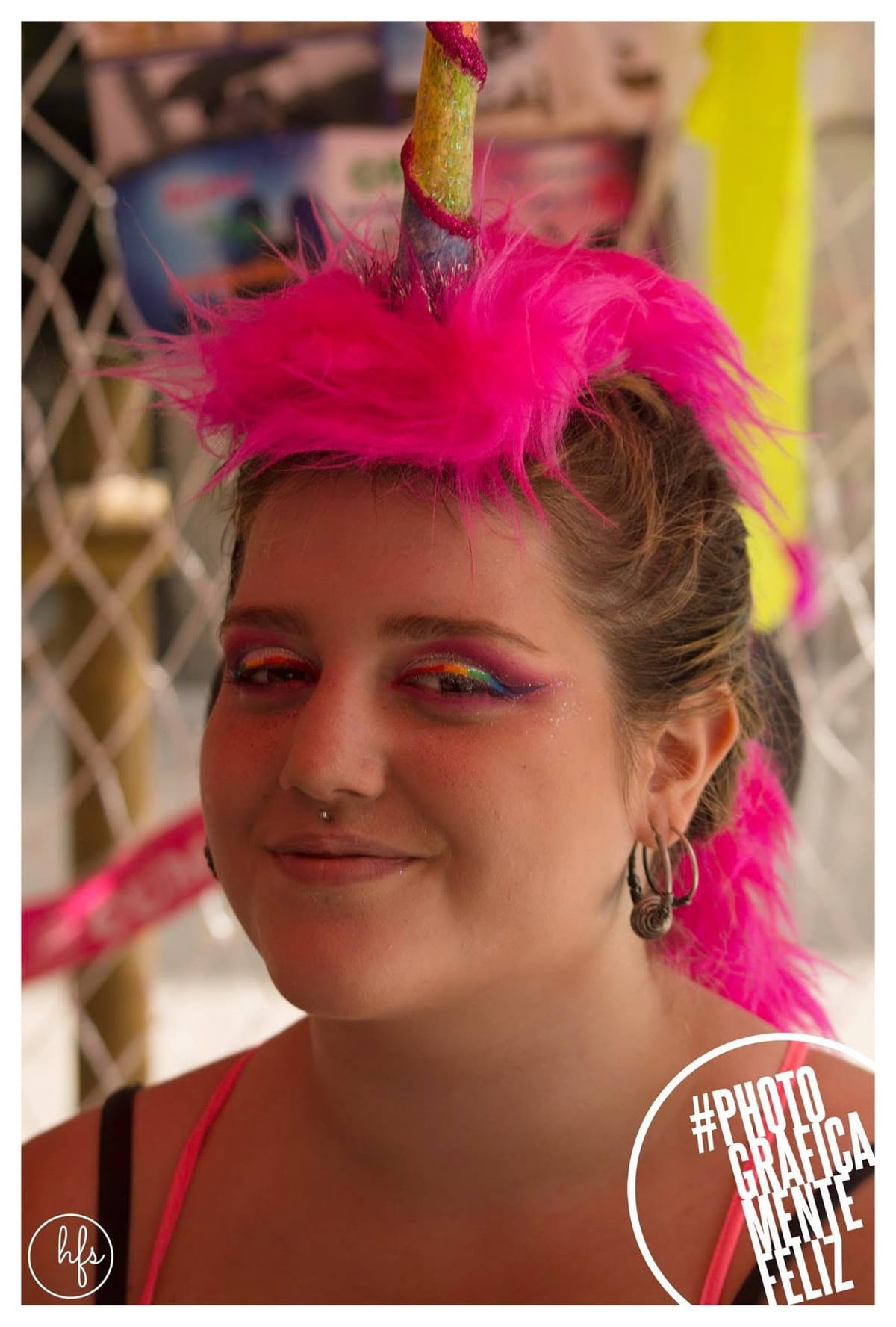 Retrato a color en primer plano de una joven con un tocado de cuerno de unicornio rosa fucsia y maquillaje de ojos arcoíris con brillantina, aros y piercing en la nariz, sonriendo levemente.