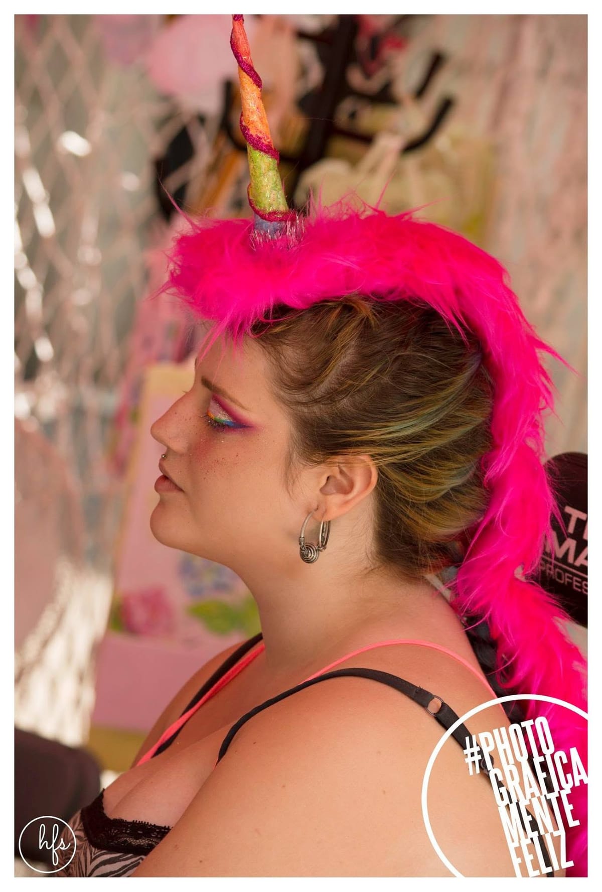Escena de backstage: una mujer de pelo castaño le aplica maquillaje en los labios a otra que ya lleva puesta la diadema de unicornio con plumas fucsia y ojos cerrados, en un espacio…