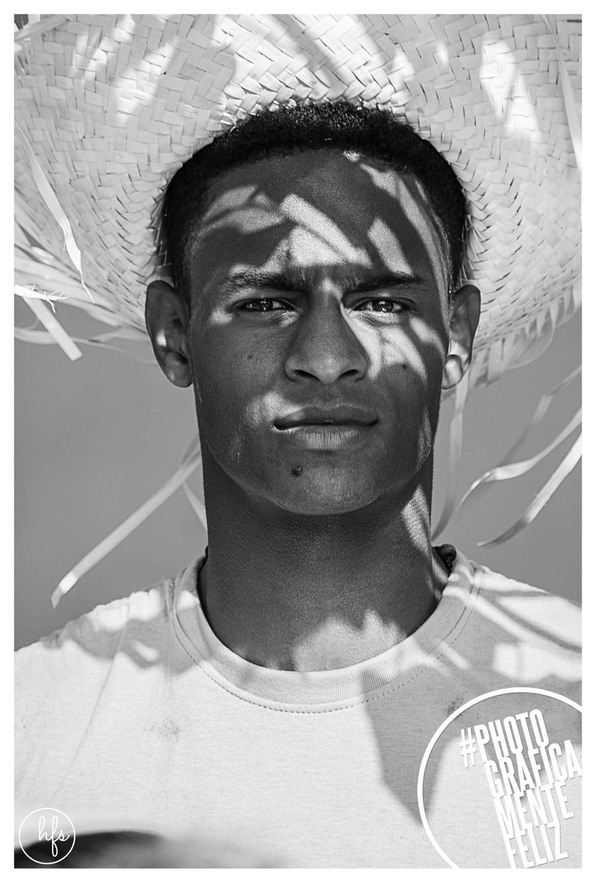 Retrato en blanco y negro de un joven con un gran sombrero de paja deshilachado, cuya sombra proyecta un patrón geométrico sobre su rostro serio; viste una remera clara, fondo neutro.
