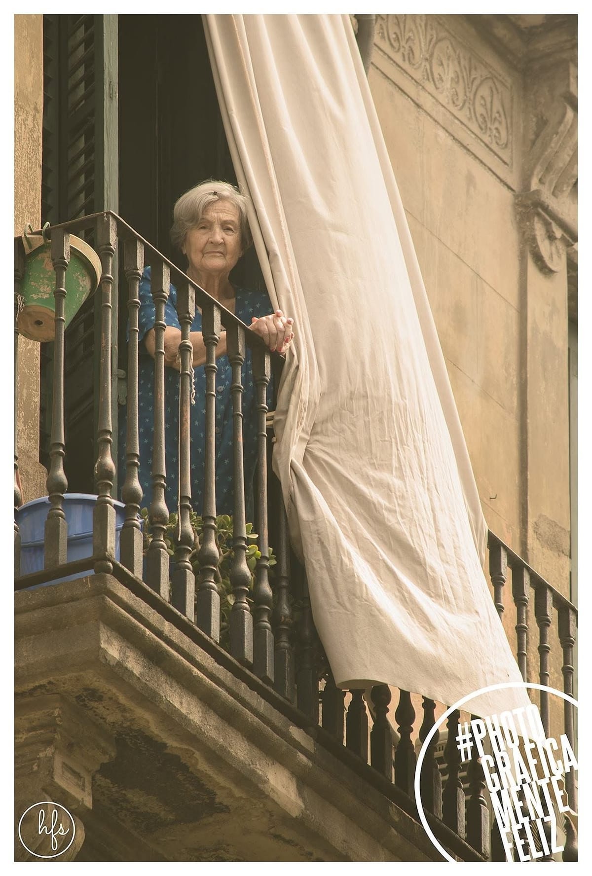 Fotografía a color de una mujer mayor de pelo blanco asomada a un balcón de hierro forjado, vestida con una bata azul de flores, sosteniendo una cortina clara que el viento agita; fachada…