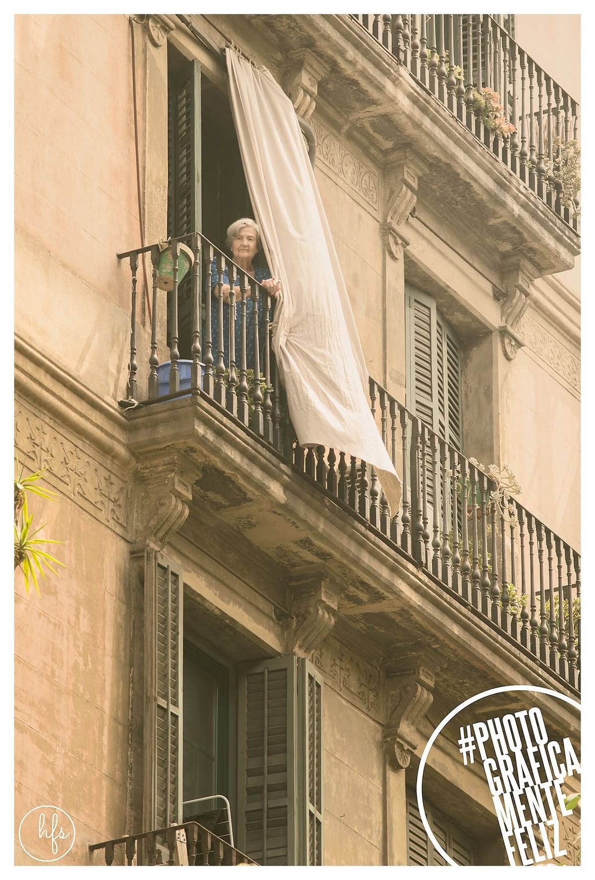 Mujer mayor de pelo blanco asomada a un balcón con una cortina clara ondeando al viento, apoyada en la baranda de hierro de un edificio histórico con fachada ornamentada, en tono sepia…