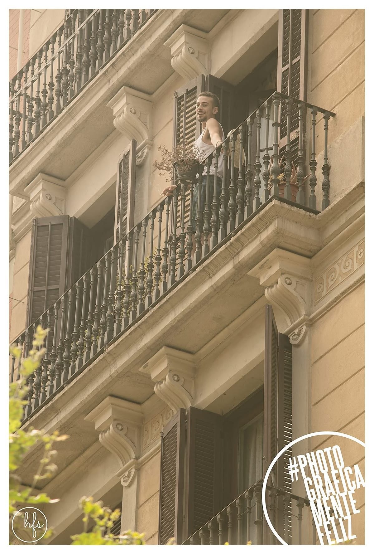 Desde la calle y en contrapicado, dos hombres se besan asomados a un balcón de un edificio modernista, uno de piel oscura vestido con túnica blanca bordada, el otro rubio y sin remera con…