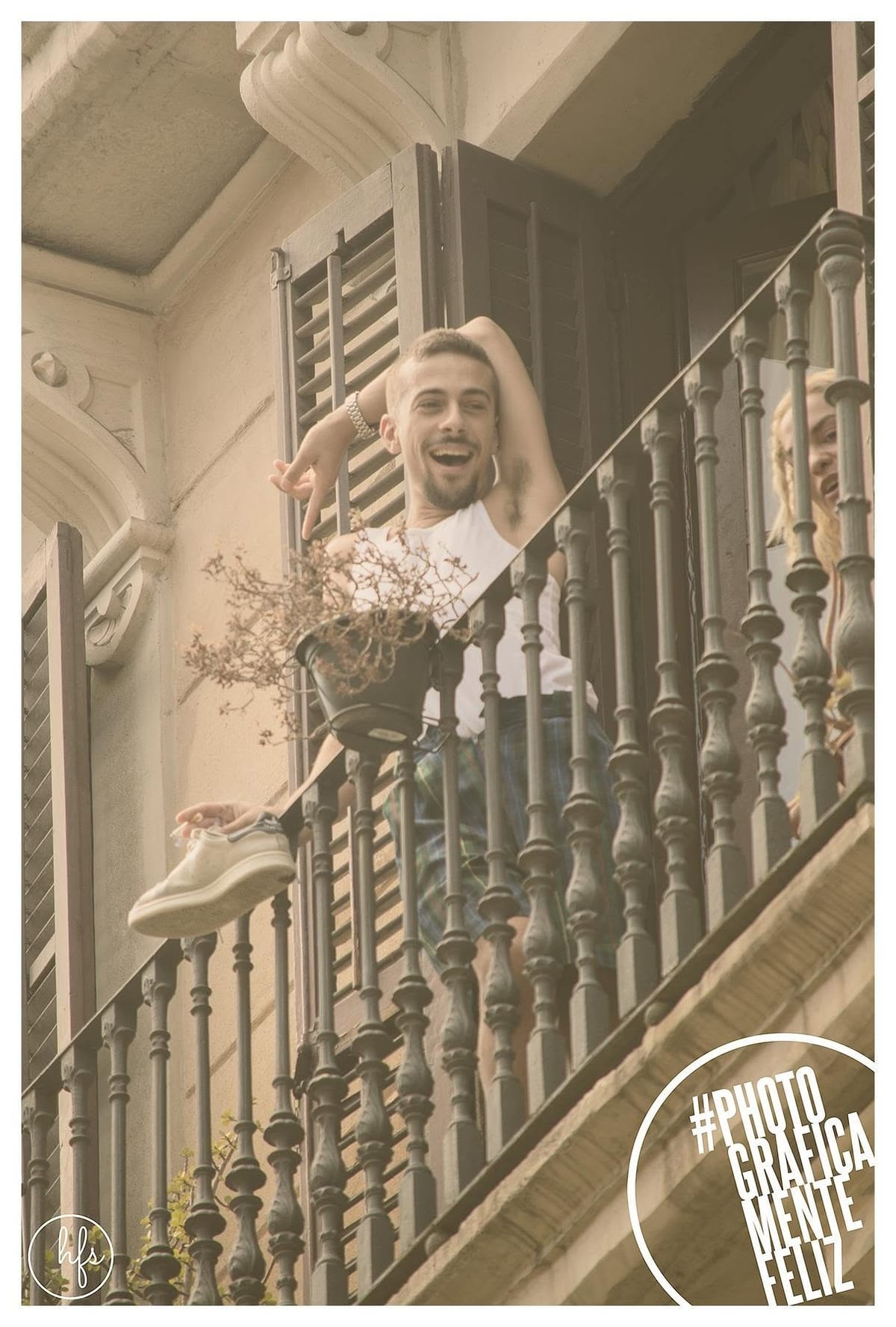 Fotografía a color en tono sepia de un joven riendo apoyado en la baranda de un balcón con postigos de madera, un brazo levantado y una zapatilla colgando del pie sobre la baranda; se…