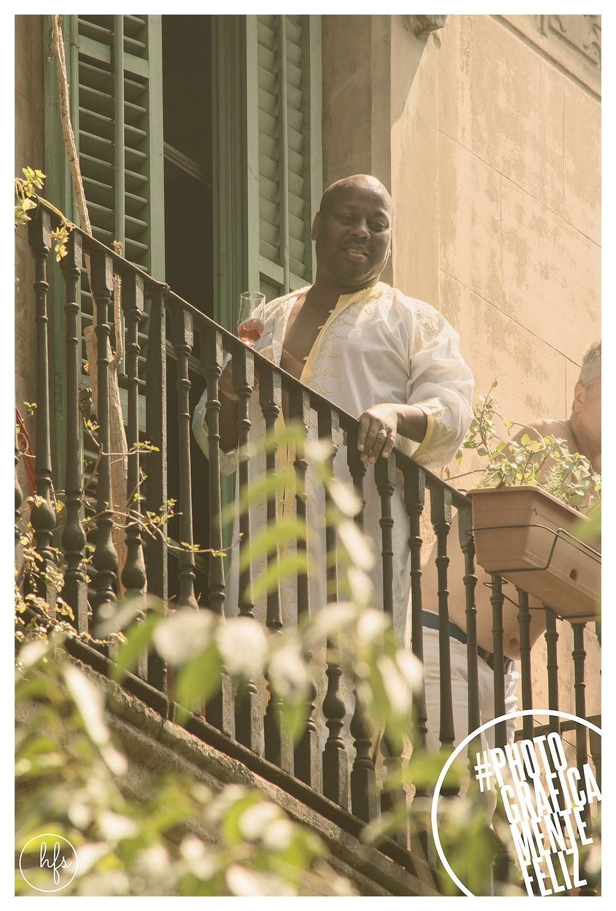 Hombre mayor con túnica blanca bordada, apoyado en la baranda de hierro de un balcón con plantas, sosteniendo una copa de vino rosado.